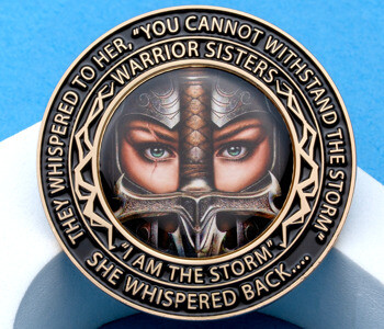 Round black and gold challenge coin featuring a woman warrior's face and sword with the text 'Warrior Sisters' and 'I am the storm'.