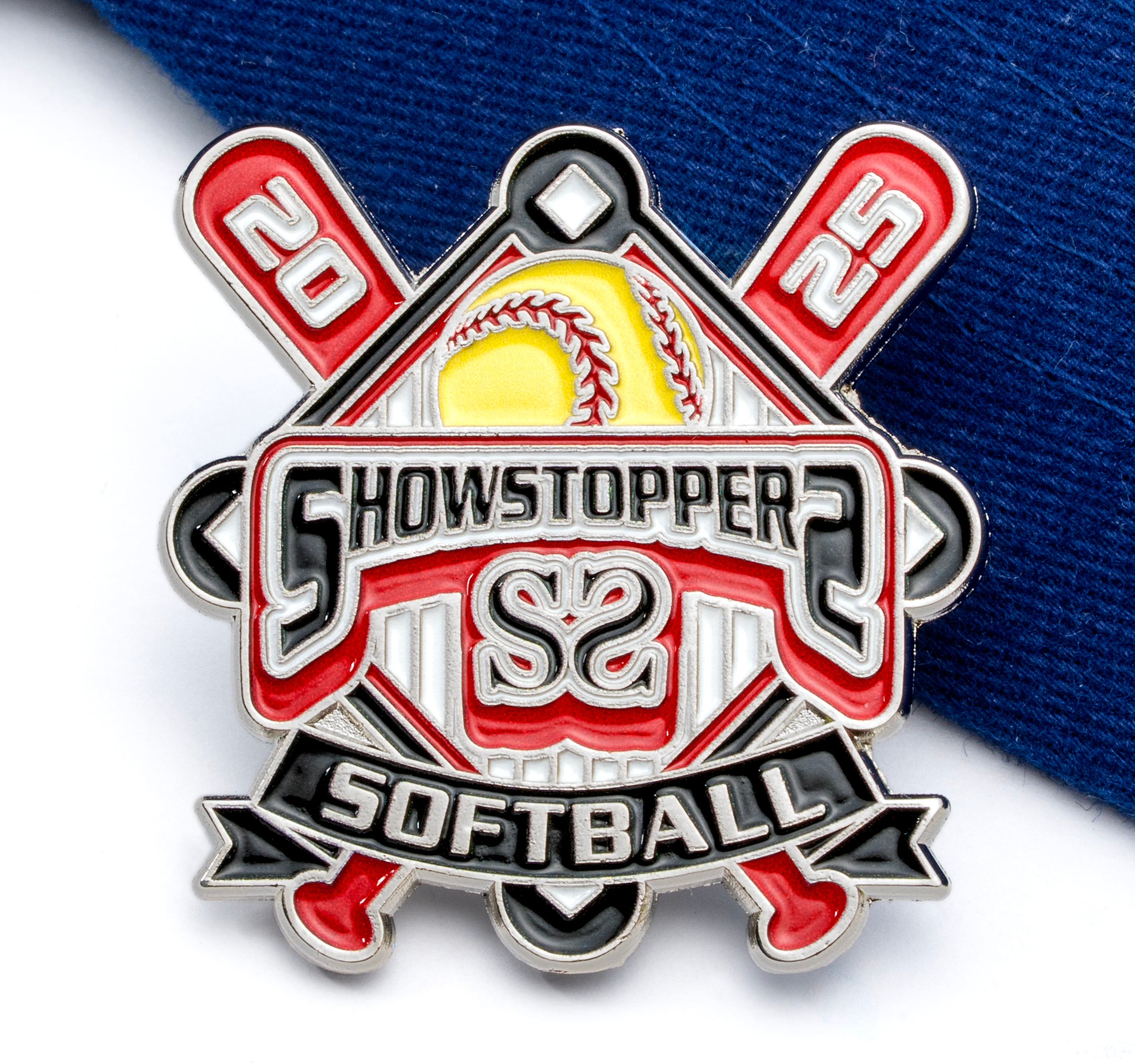 Soft enamel lapel pin for Showstopper Softball 2025 featuring crossed bats, a yellow softball, and red, black, and white accents on a blue fabric background.