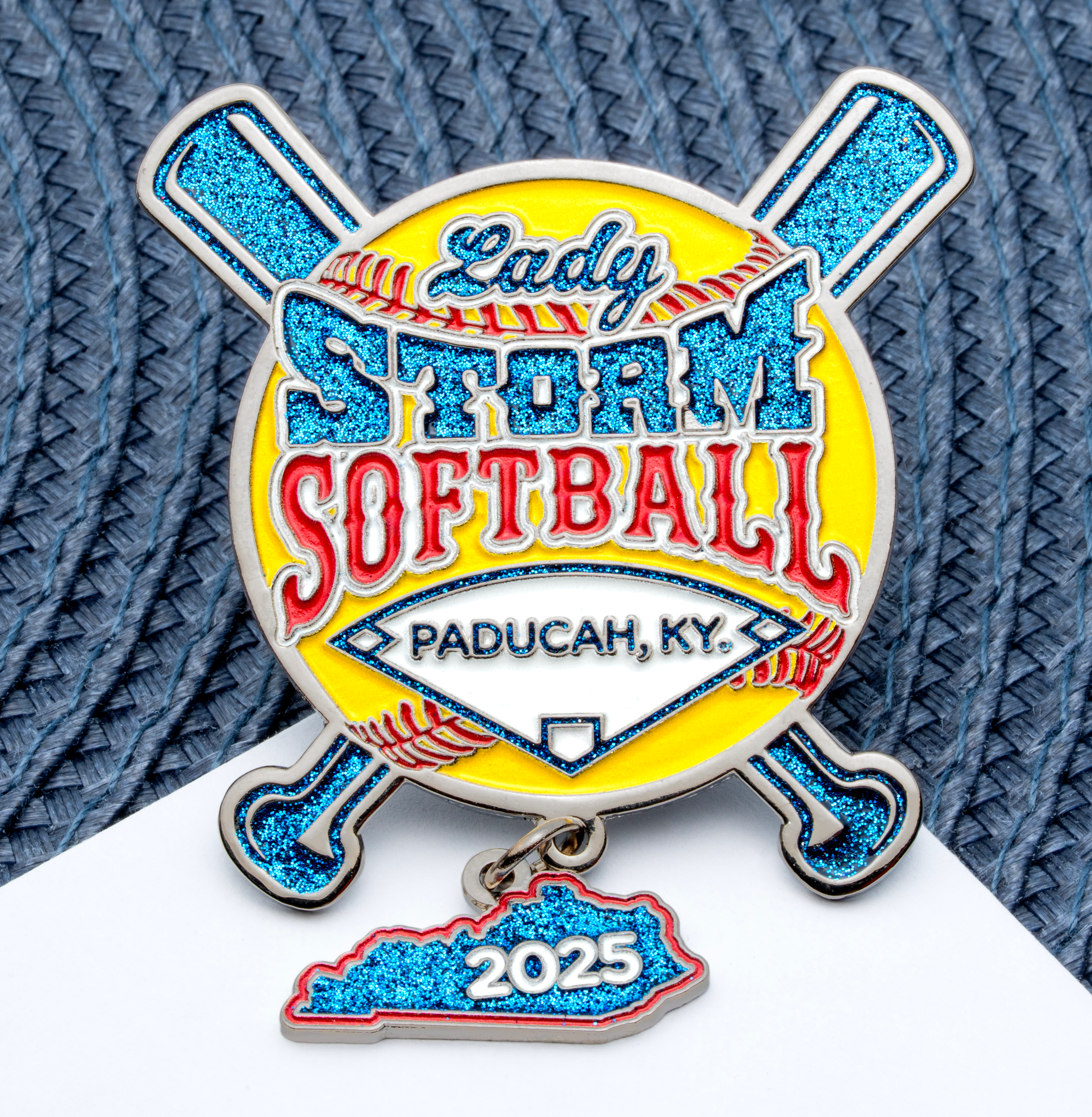 Custom softball lapel pin featuring crossed bats, a yellow ball, blue glitter text, and a Kentucky state-shaped charm dated 2025.
