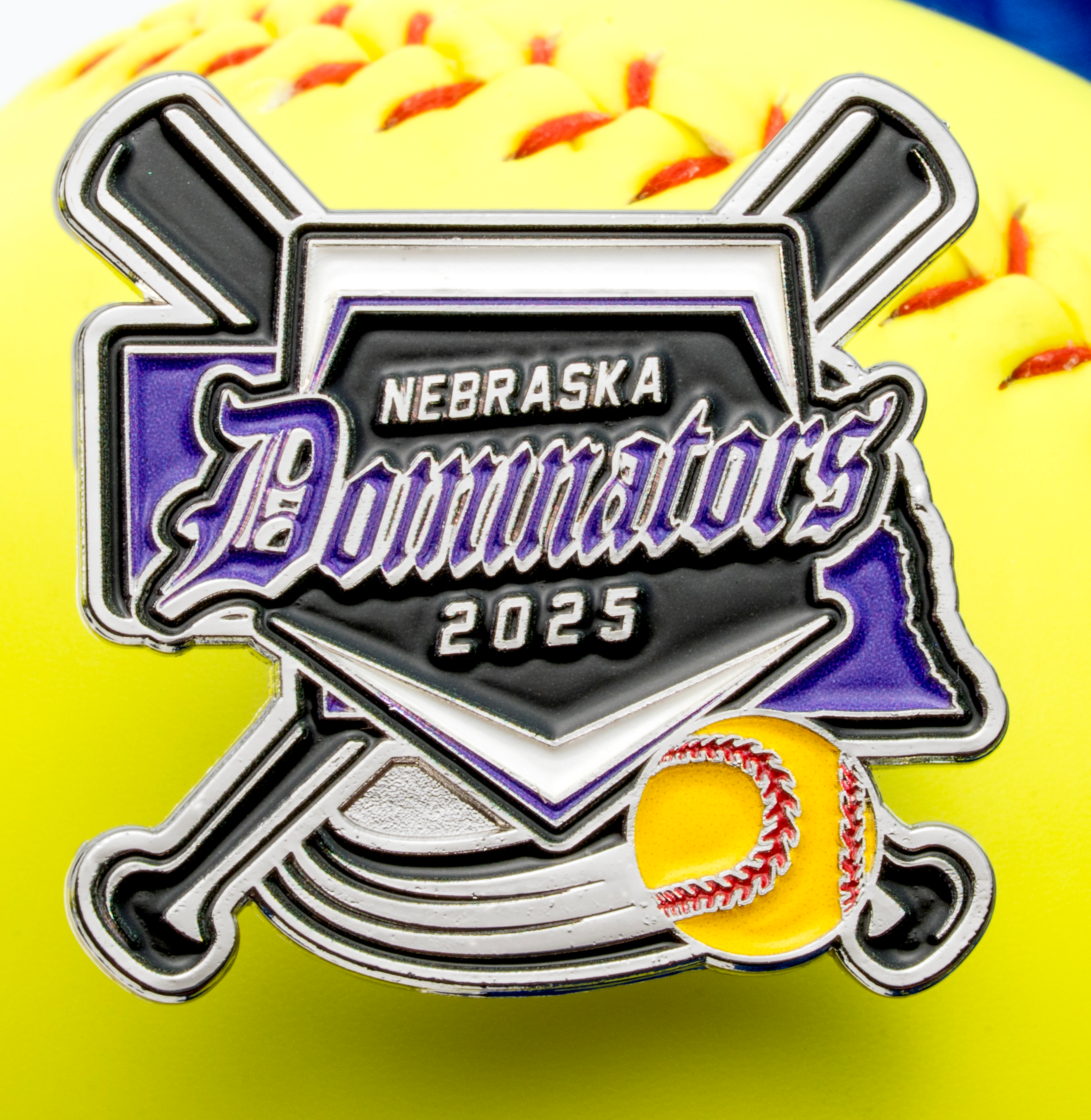 Softball lapel pin featuring Nebraska Dominators 2025 text, crossed bats, a home plate, and a yellow softball over a larger yellow softball background.