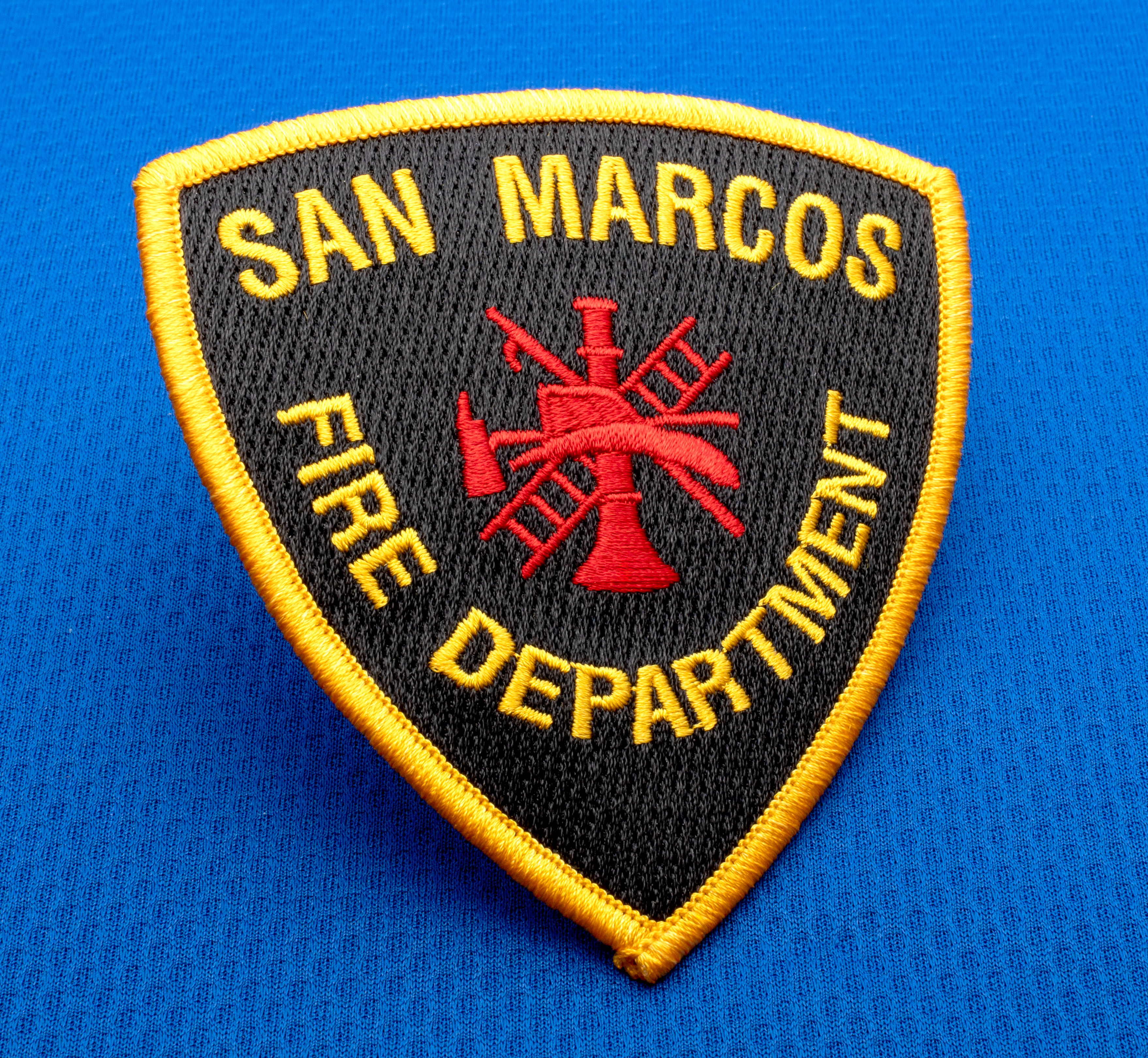 map[name:Shield-shaped embroidered patch for San Marcos Fire Department featuring a red scramble icon on a black background with a gold border. naturalSort:Shield-shaped embroidered patch for San Marcos Fire Department featuring a red scramble icon on a black background with a gold border.]