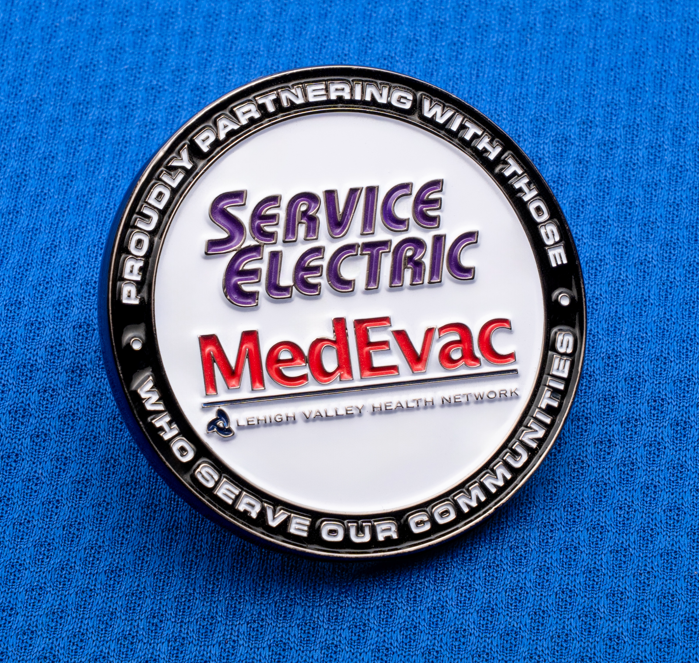 map[name:Circular lapel pin featuring Service Electric and MedEvac logos on a white background with a black border on blue textured fabric. naturalSort:Circular lapel pin featuring Service Electric and MedEvac logos on a white background with a black border on blue textured fabric.]