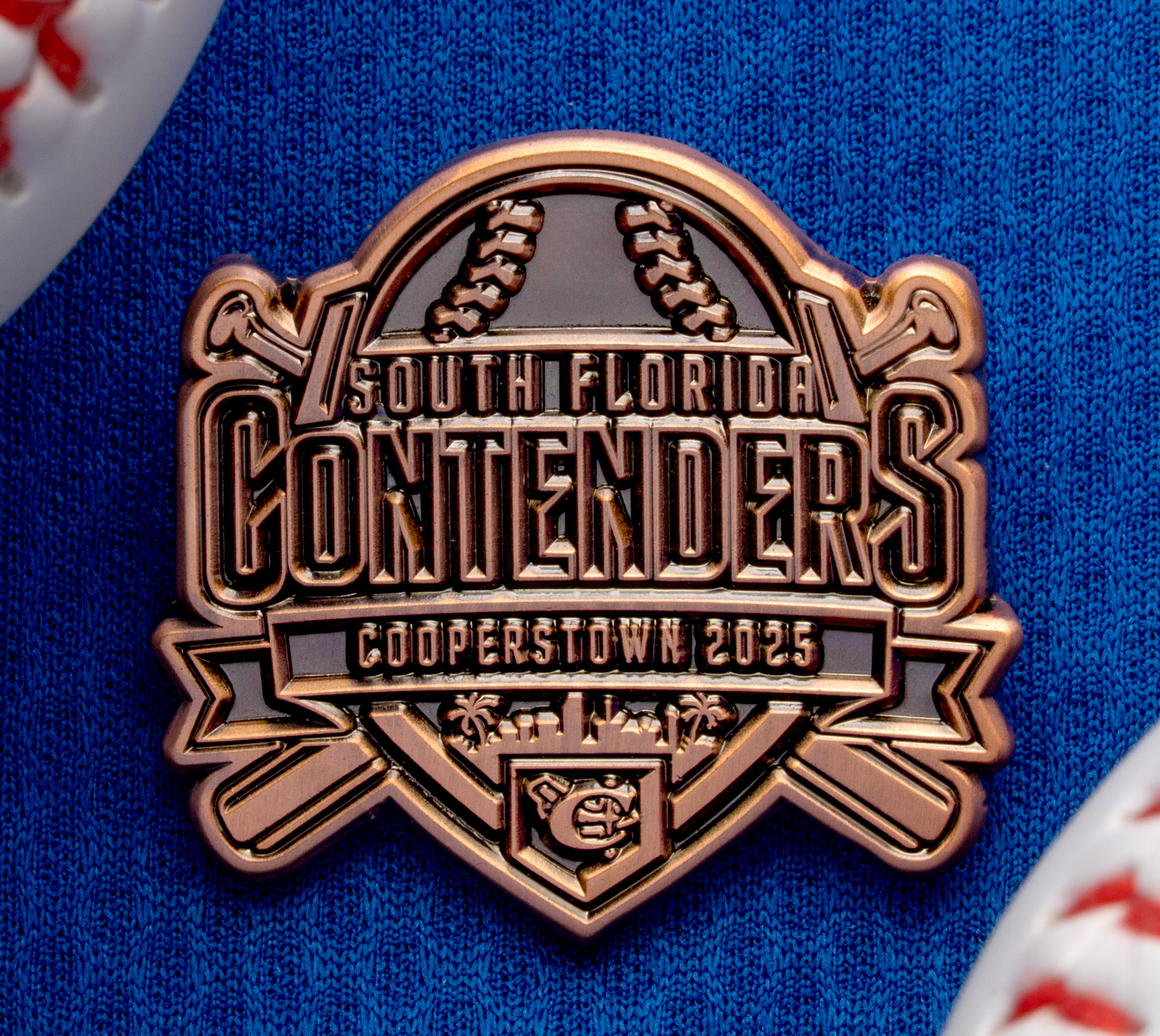 Bronze-colored South Florida Contenders Cooperstown 2025 baseball trading pin between two baseballs on a blue textured background.