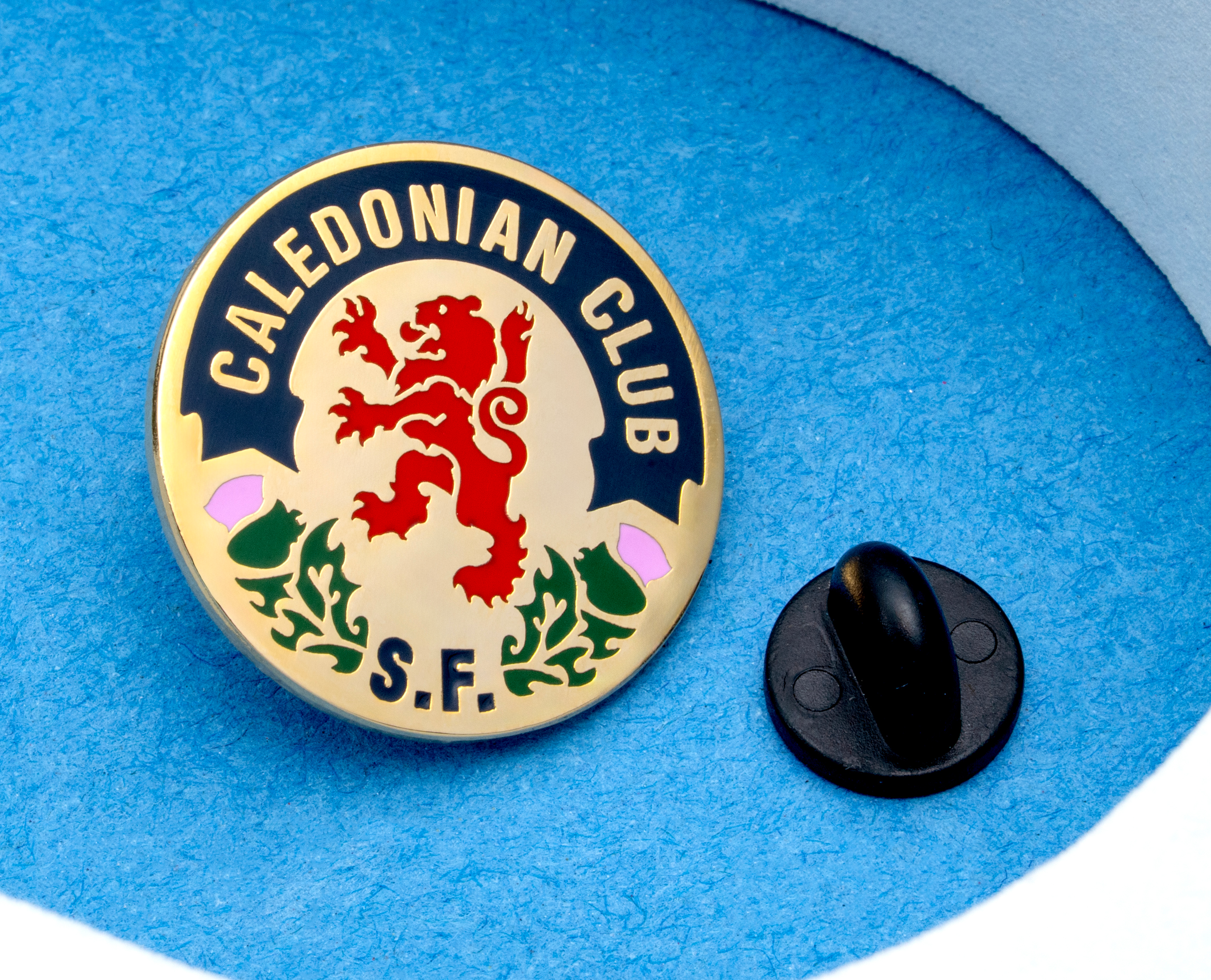 Circular gold enamel lapel pin for Caledonian Club S.F. featuring a red lion rampant and thistle design on a blue background.