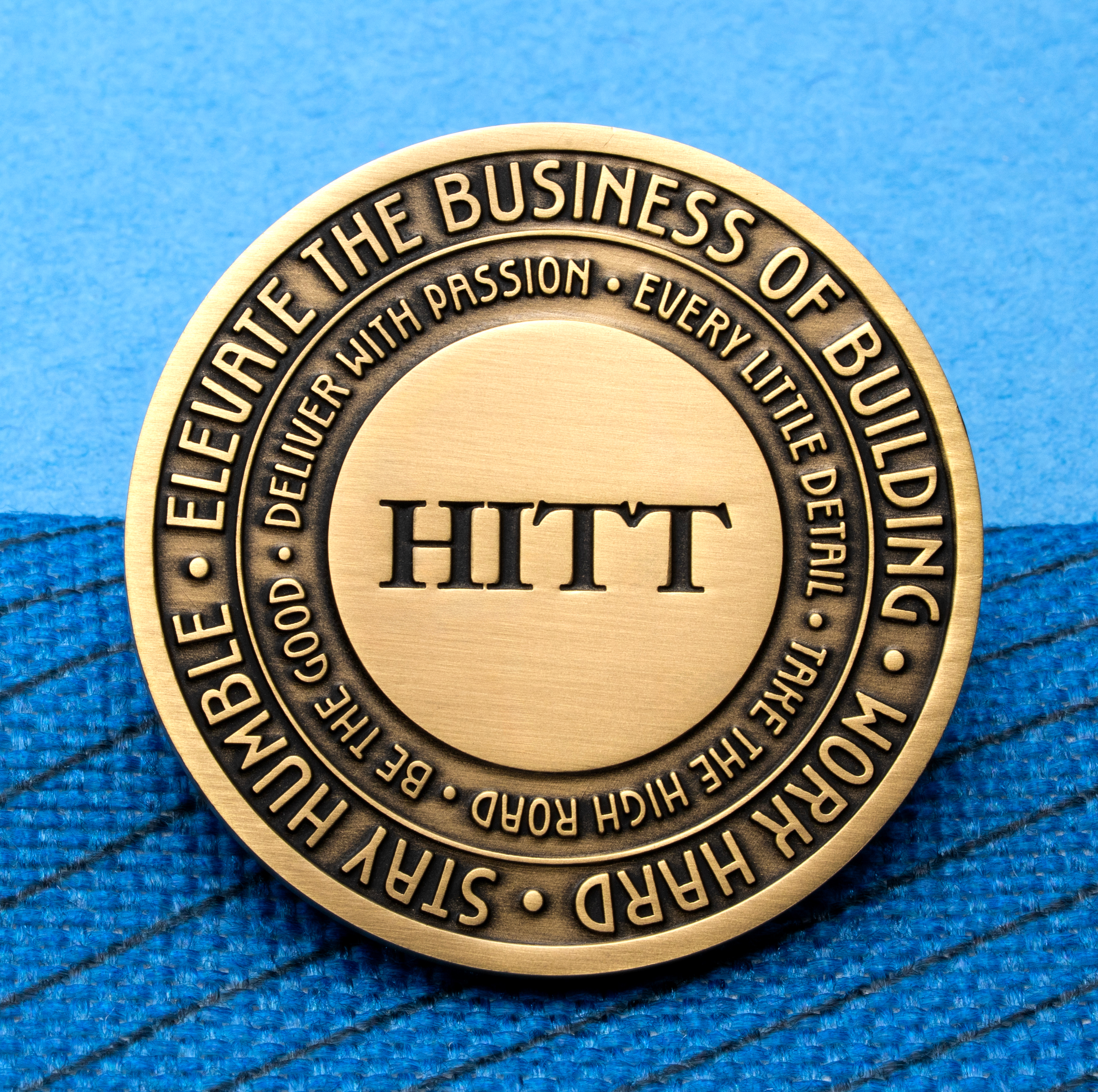 Antique brass challenge coin featuring HITT logo and concentric circles of engraved text on a blue background.
