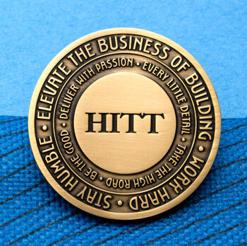 Antique brass challenge coin featuring HITT logo and concentric circles of engraved text on a blue background.