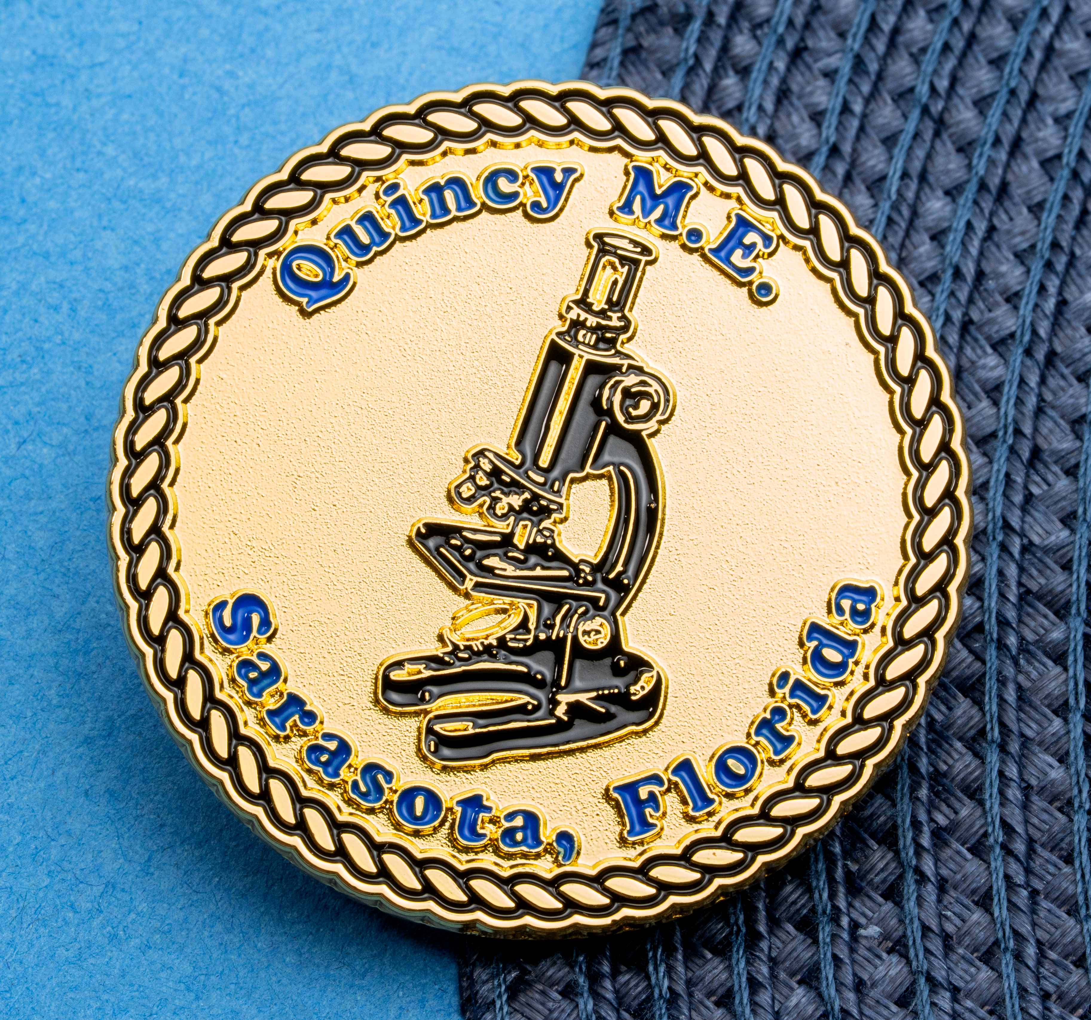 Gold circular lapel pin with a rope border featuring a black and white microscope illustration and blue text for Quincy M.E. Sarasota, Florida.