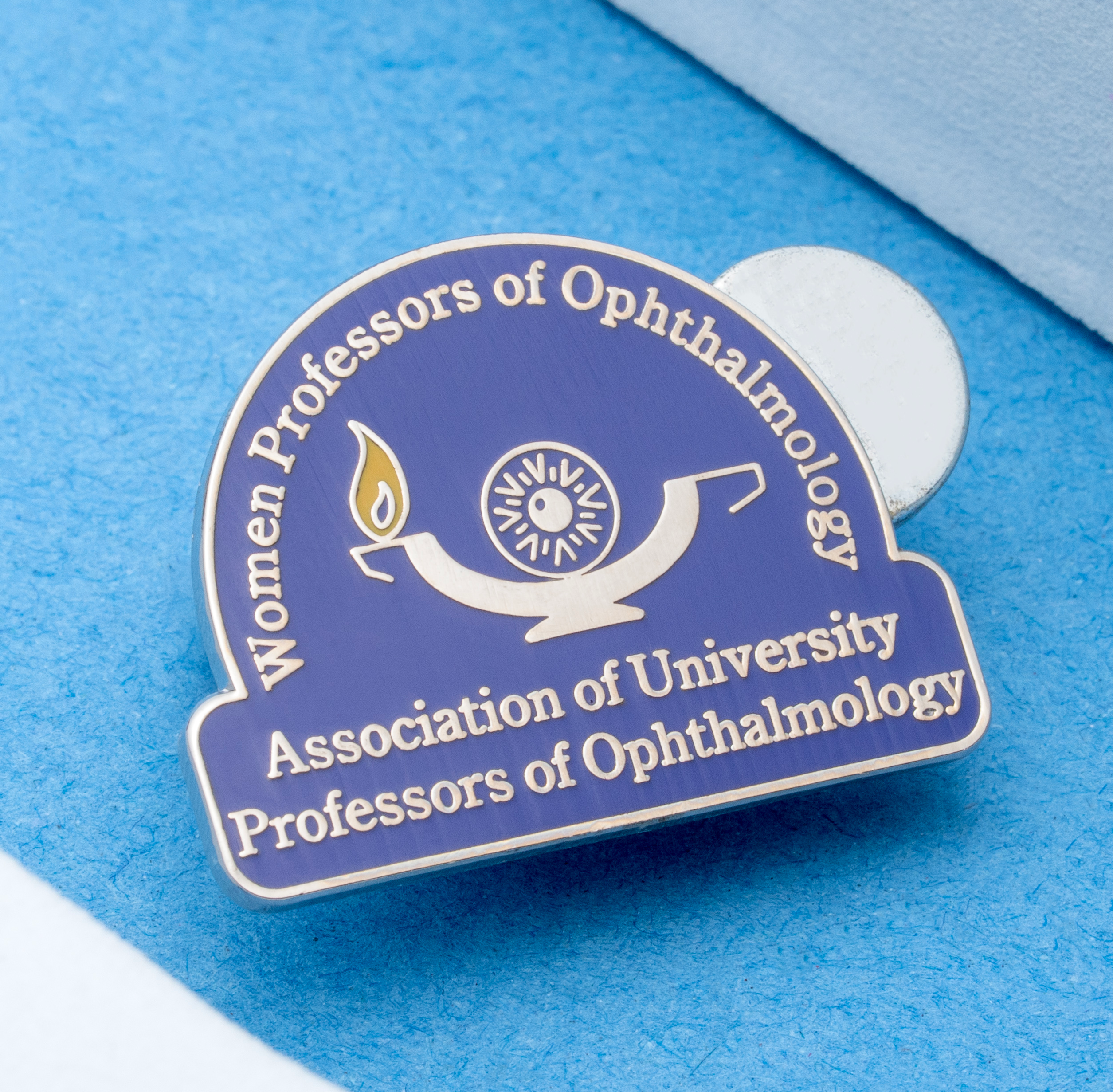 map[name:Blue and silver lapel pin for the Women Professors of Ophthalmology, featuring an oil lamp logo and gold text on a blue background. naturalSort:Blue and silver lapel pin for the Women Professors of Ophthalmology, featuring an oil lamp logo and gold text on a blue background.]