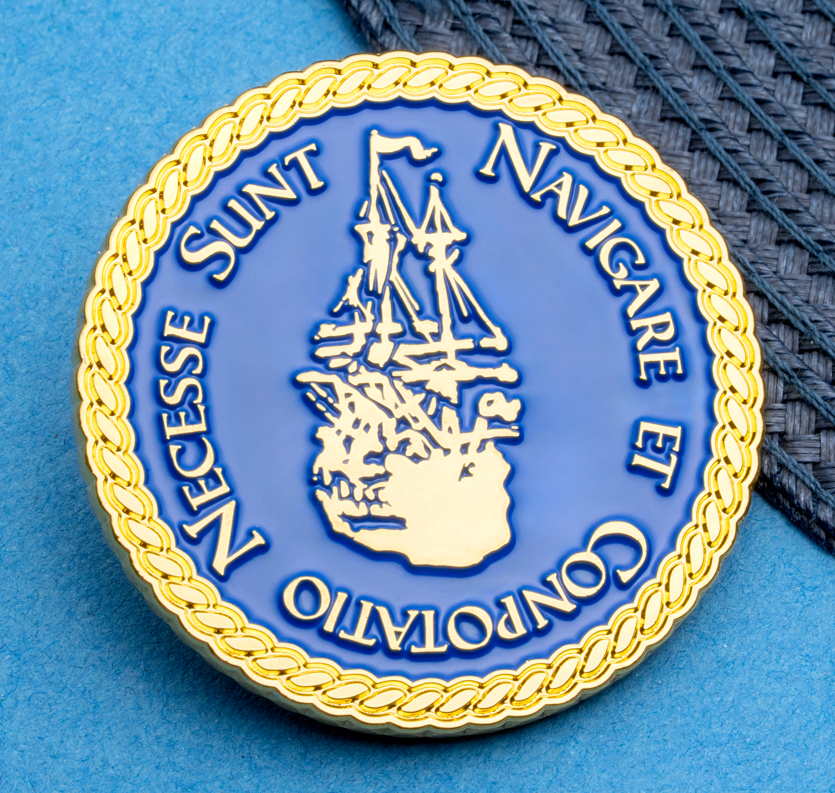 Circular blue and gold enamel coin featuring a sailing ship and the Latin text Navigare Et Conpotatio Necesse Sunt with a rope border.