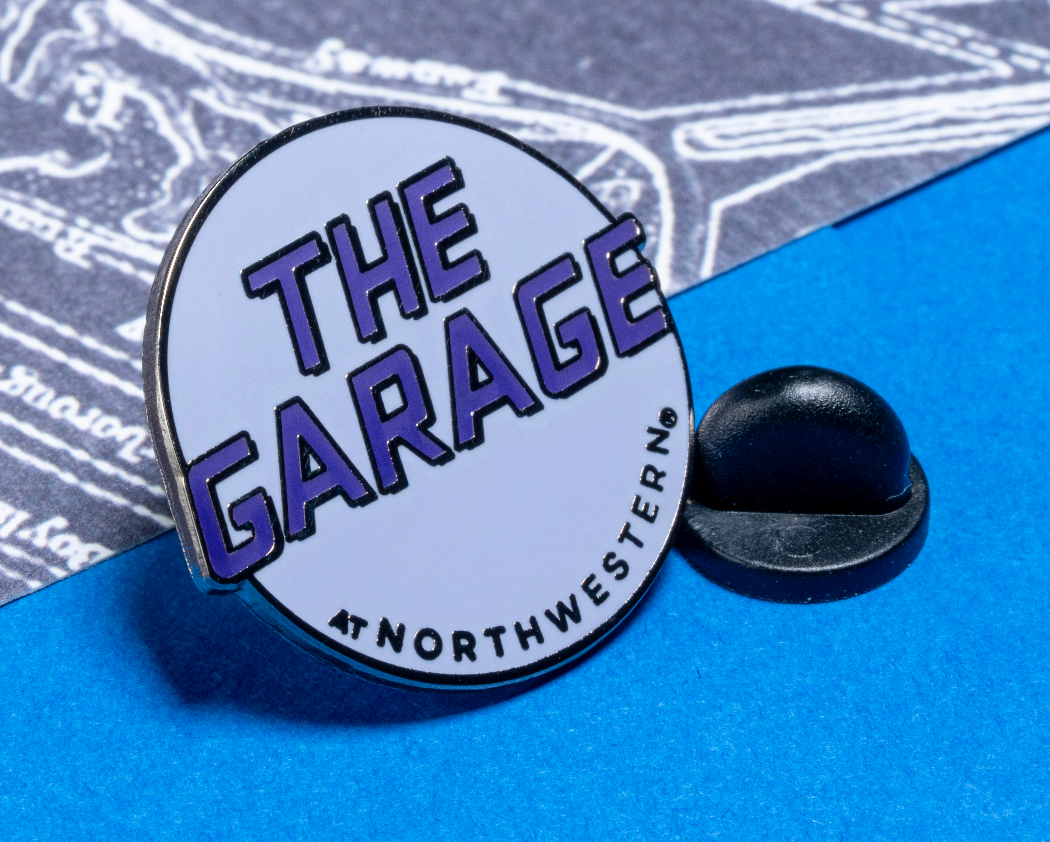 map[name:A circular enamel lapel pin with 'The Garage' in purple text and 'At Northwestern' in black text on a white background, pictured with a black rubber clutch. naturalSort:A circular enamel lapel pin with 'The Garage' in purple text and 'At Northwestern' in black text on a white background, pictured with a black rubber clutch.]