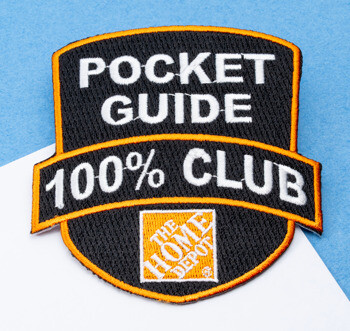 Embroidered shield-shaped patch with black background, orange borders, and white text reading Pocket Guide 100% Club above The Home Depot logo.