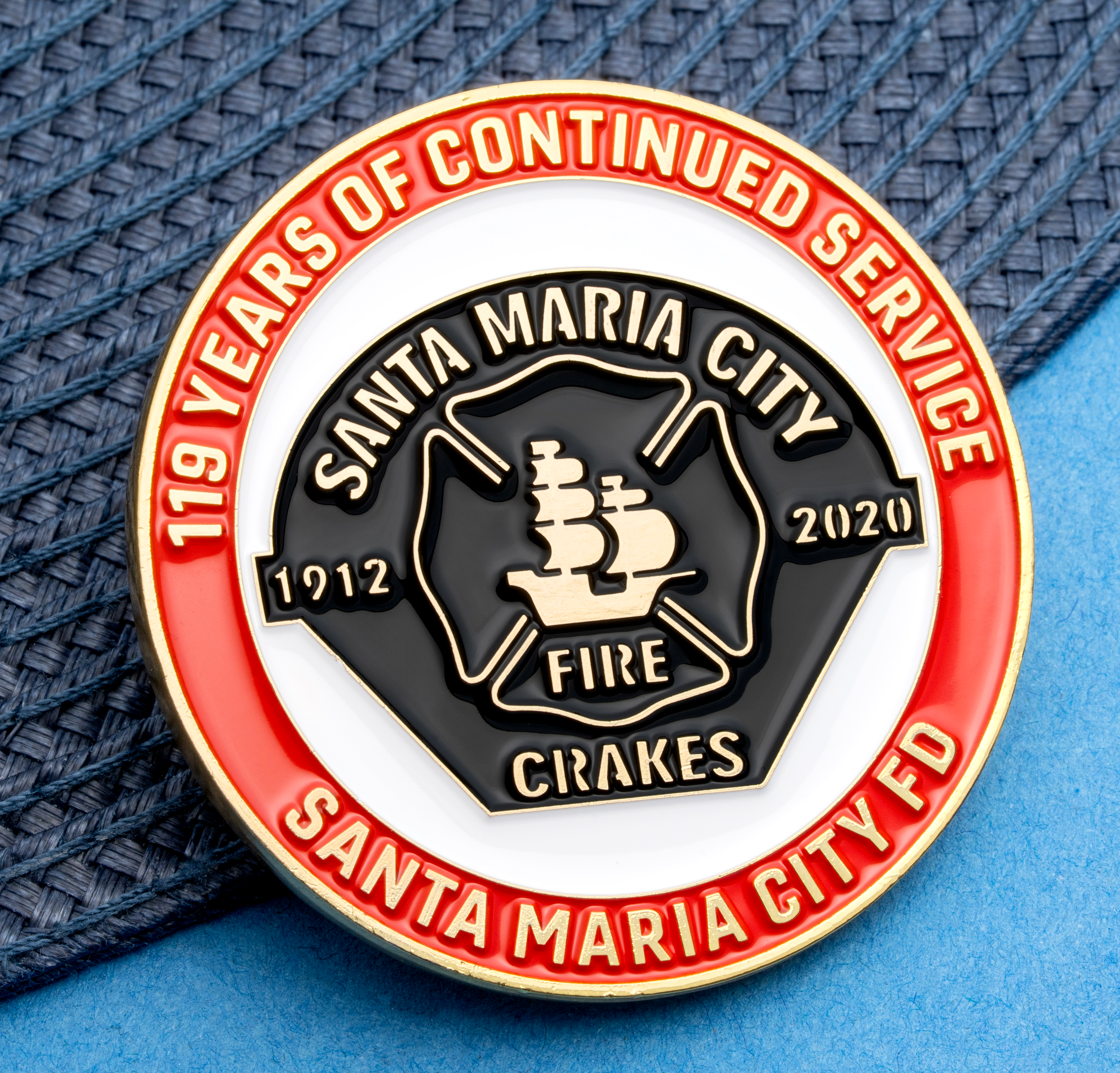 Circular enamel challenge coin for Santa Maria City Fire Department with a black shield, sailing ship, and red border on a blue textured background.