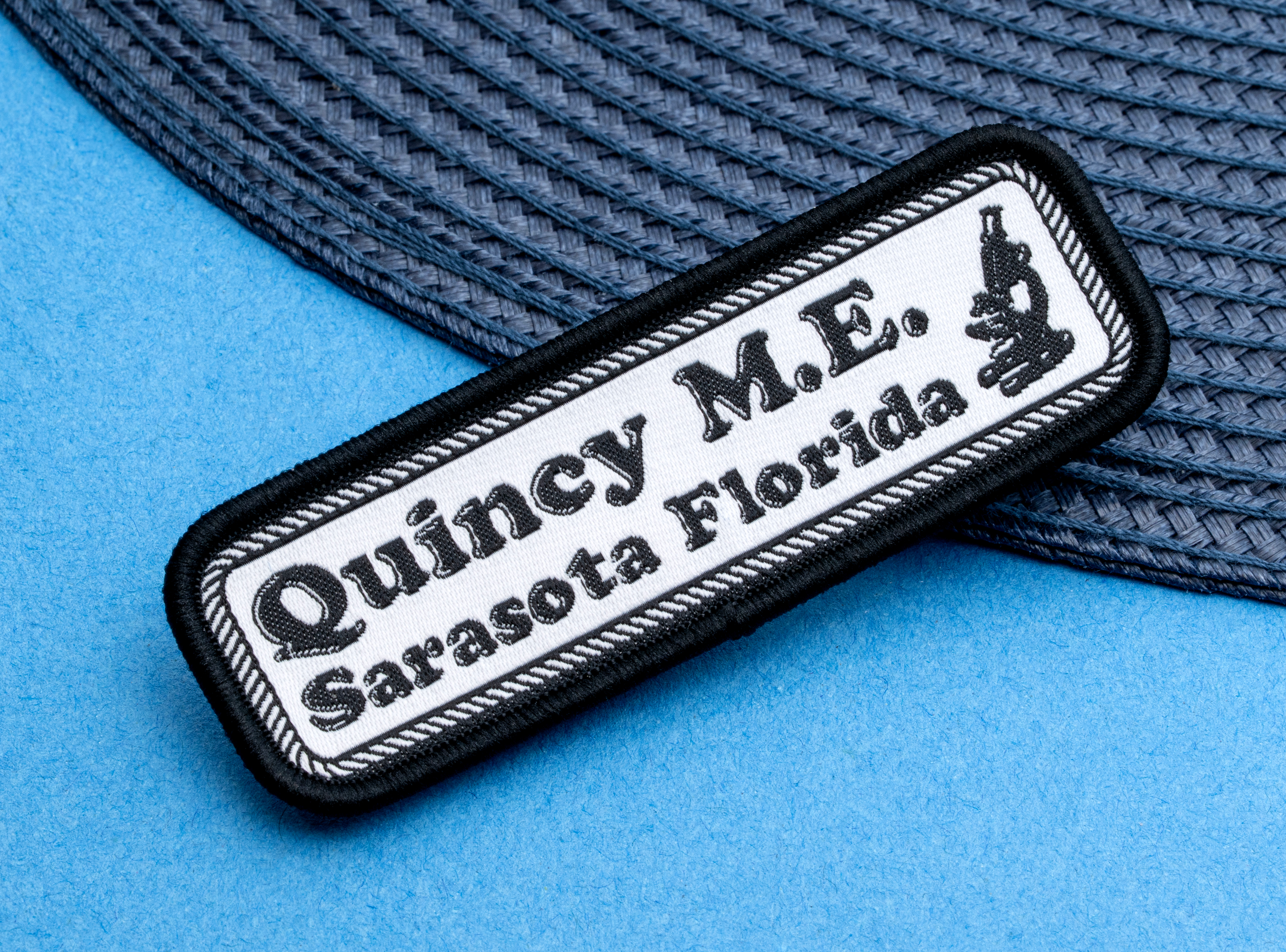 map[name:Rectangular embroidered patch with black text reading Quincy M.E. Sarasota Florida and a microscope icon on a white background with black border. naturalSort:Rectangular embroidered patch with black text reading Quincy M.E. Sarasota Florida and a microscope icon on a white background with black border.]