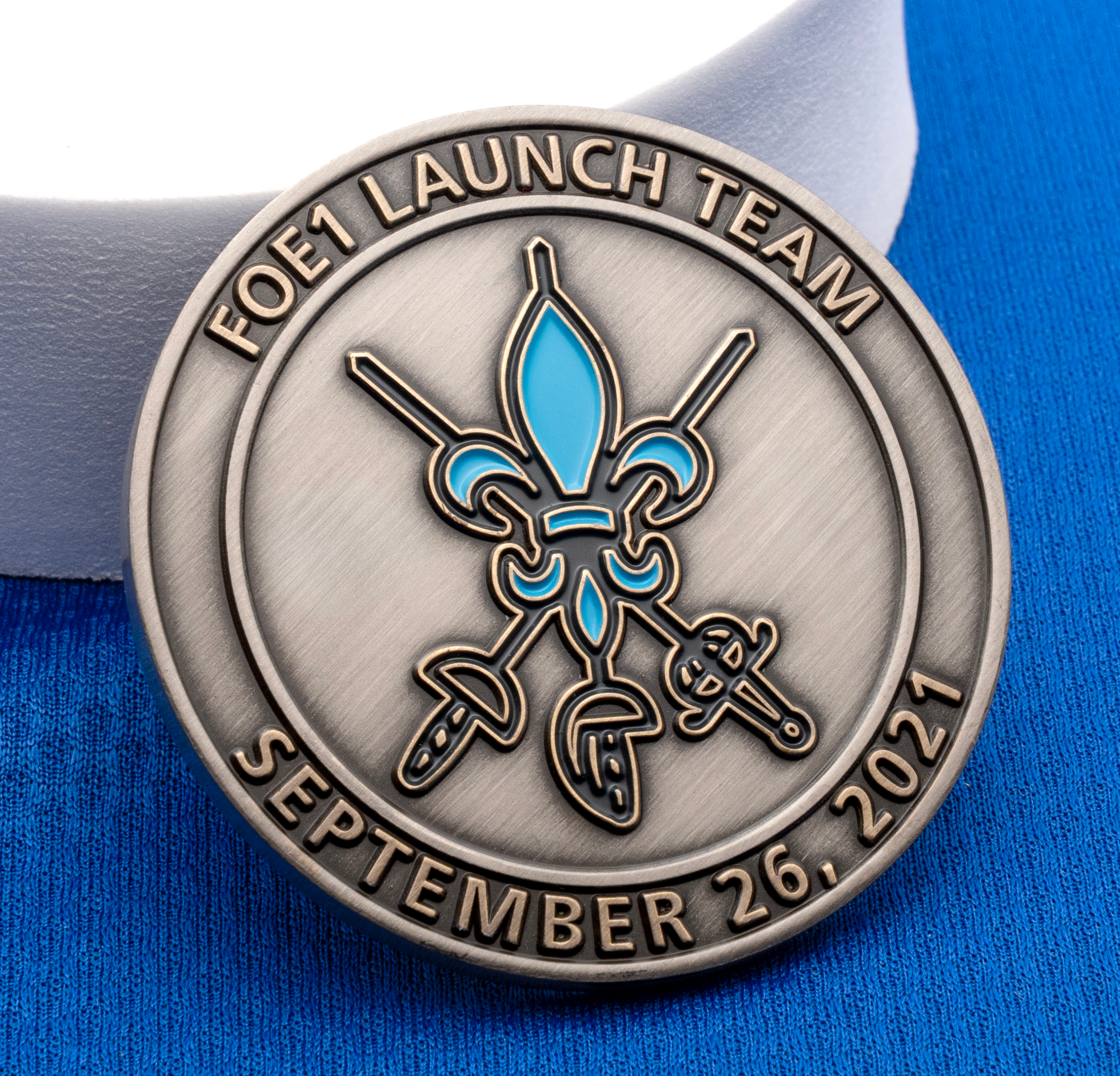 Antiqued silver challenge coin featuring a blue fleur-de-lis and crossed swords for the FOE1 Launch Team dated September 26, 2021.