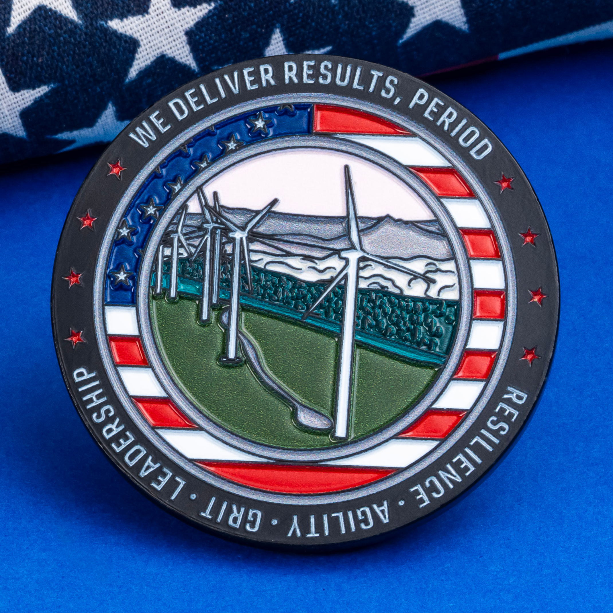 Circular challenge coin featuring a wind farm landscape and American flag border, resting on a flag backdrop.