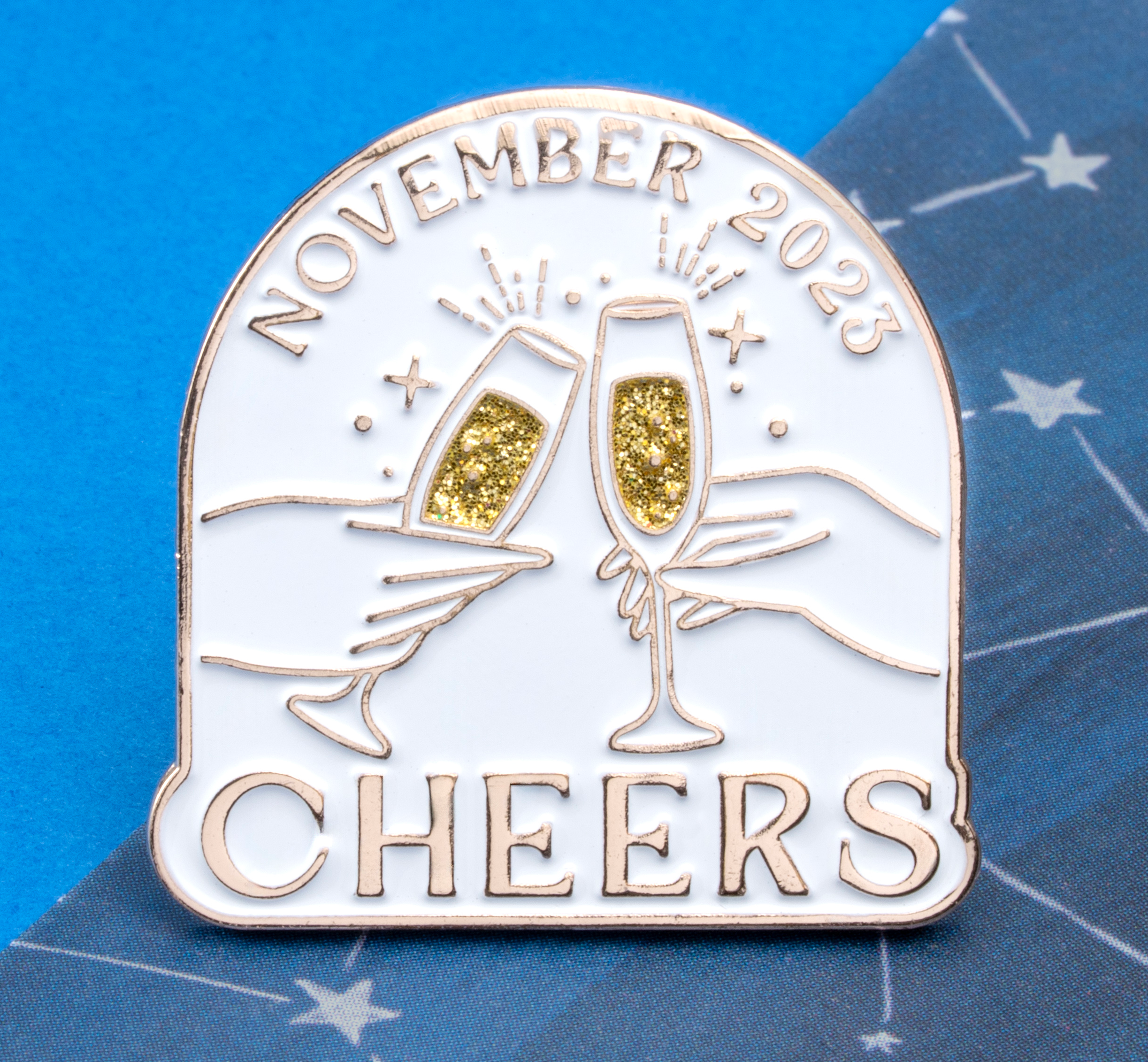 Arched white lapel pin with gold trim featuring two hands clinking champagne glasses with gold glitter, 'November 2023' at the top, and 'CHEERS' at the bottom.