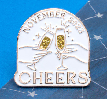 Arched white lapel pin with gold trim featuring two hands clinking champagne glasses with gold glitter, 'November 2023' at the top, and 'CHEERS' at the bottom.