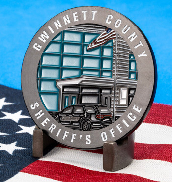 Gwinnett County Sheriff's Office challenge coin featuring a patrol vehicle, building, and American flag on a stand over a flag background.