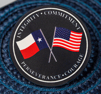 Circular embroidered patch featuring crossed Texas and United States flags with the words Integrity, Commitment, Perseverance, and Courage.