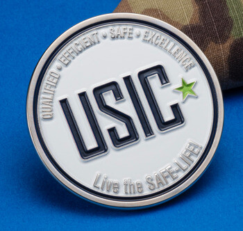 Circular USIC challenge coin featuring white enamel, black lettering, and a green star, set against a blue background and camouflage fabric.