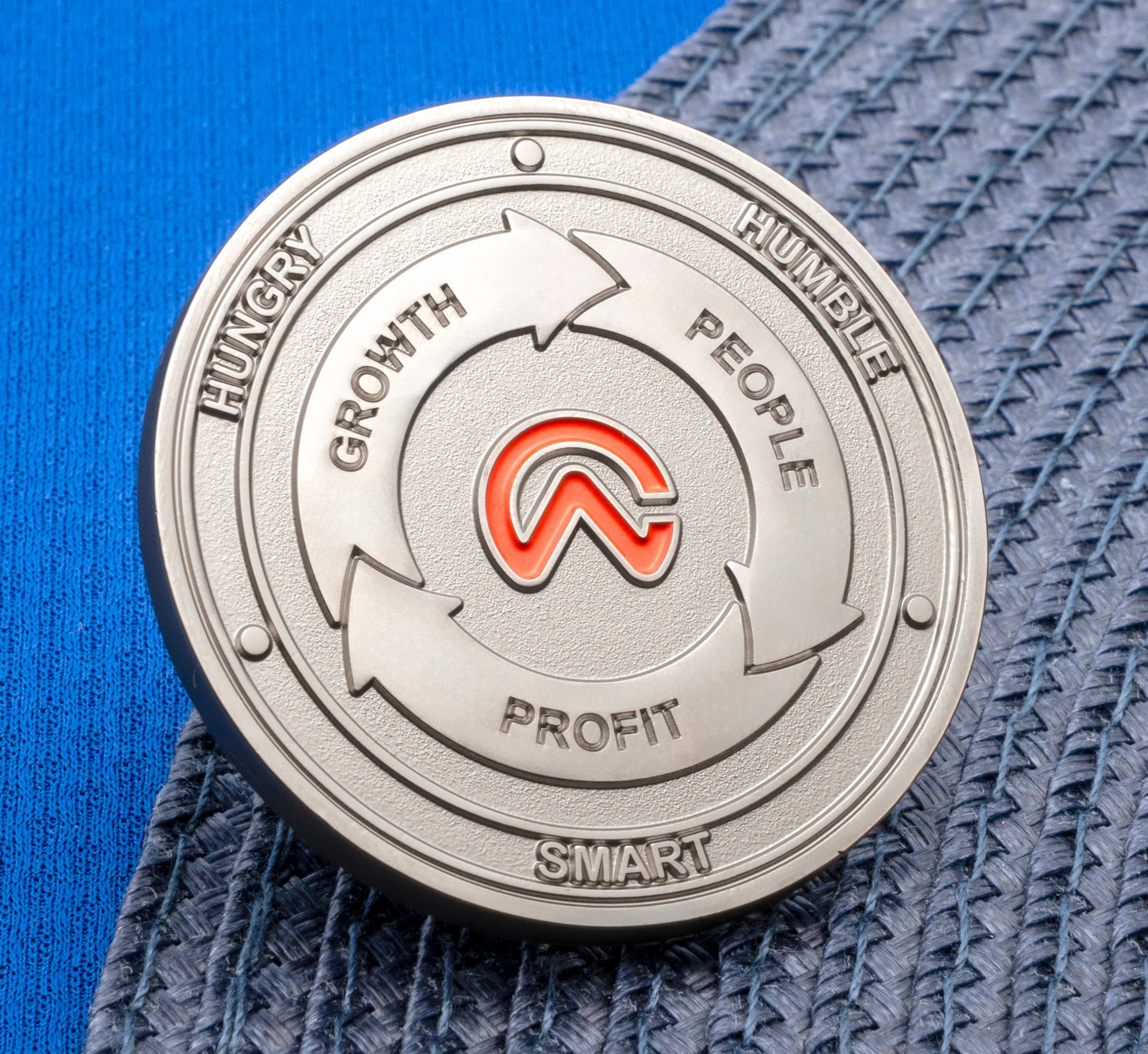 Silver challenge coin with orange logo and inscribed words Hungry, Humble, Smart, Growth, People, and Profit.
