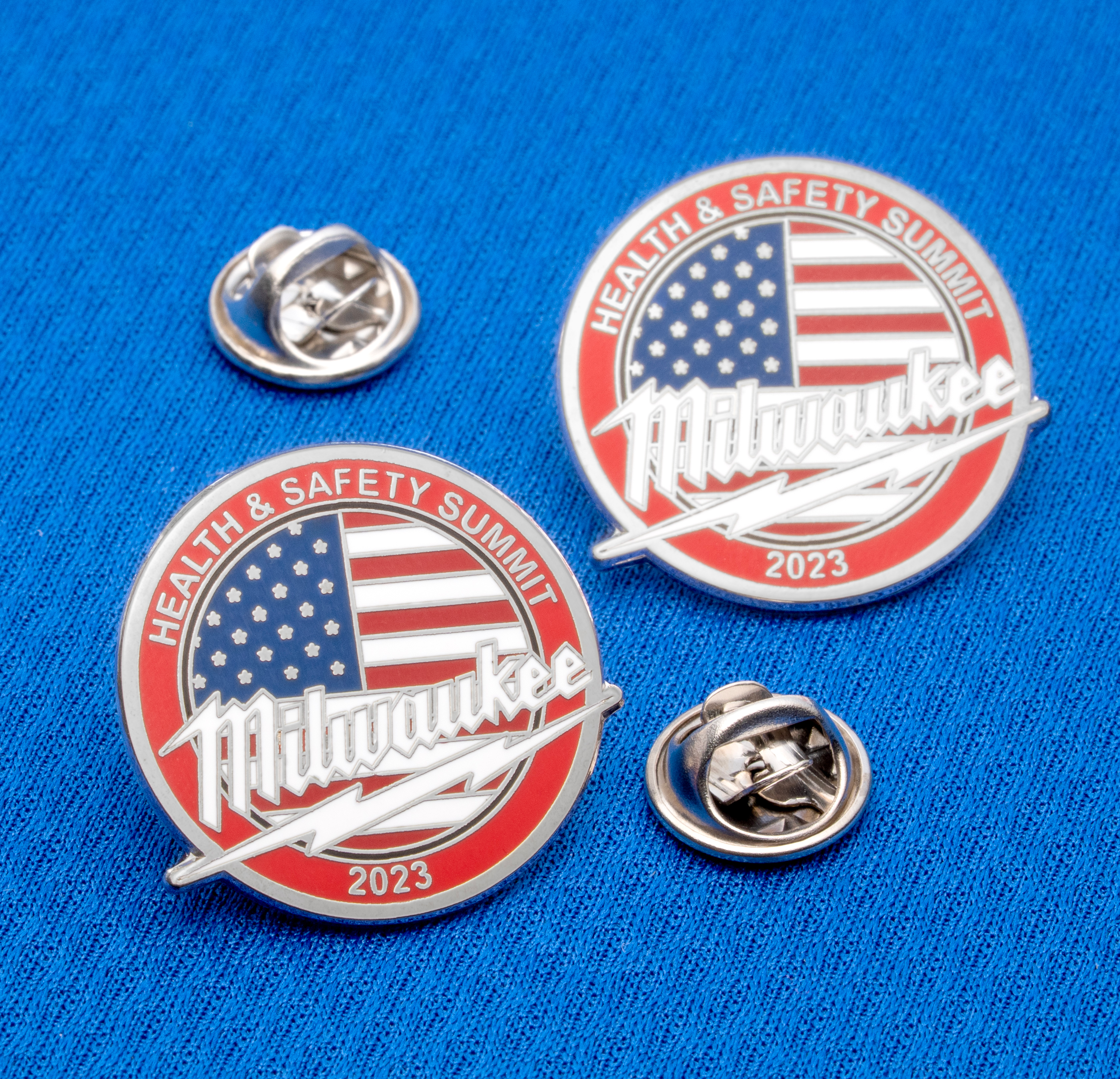 Round lapel pins featuring the American flag, Milwaukee script, and Health and Safety Summit 2023 text on a blue background.