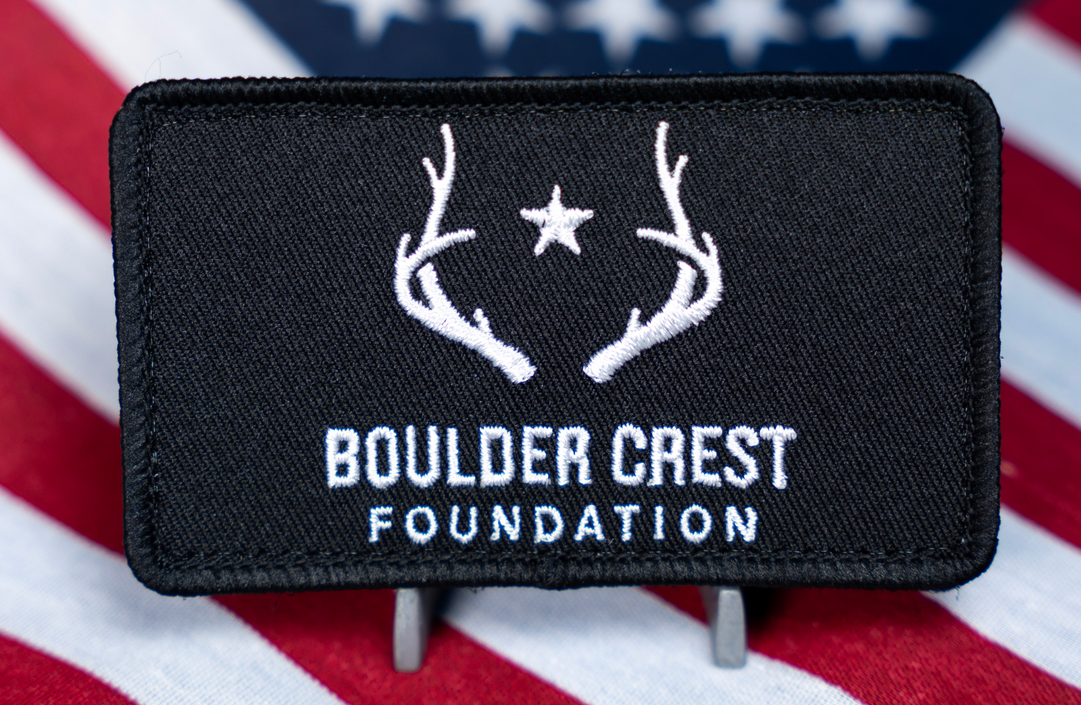 Black rectangular embroidered patch featuring white antlers and a star above the Boulder Crest Foundation text, set against a background of the American flag.