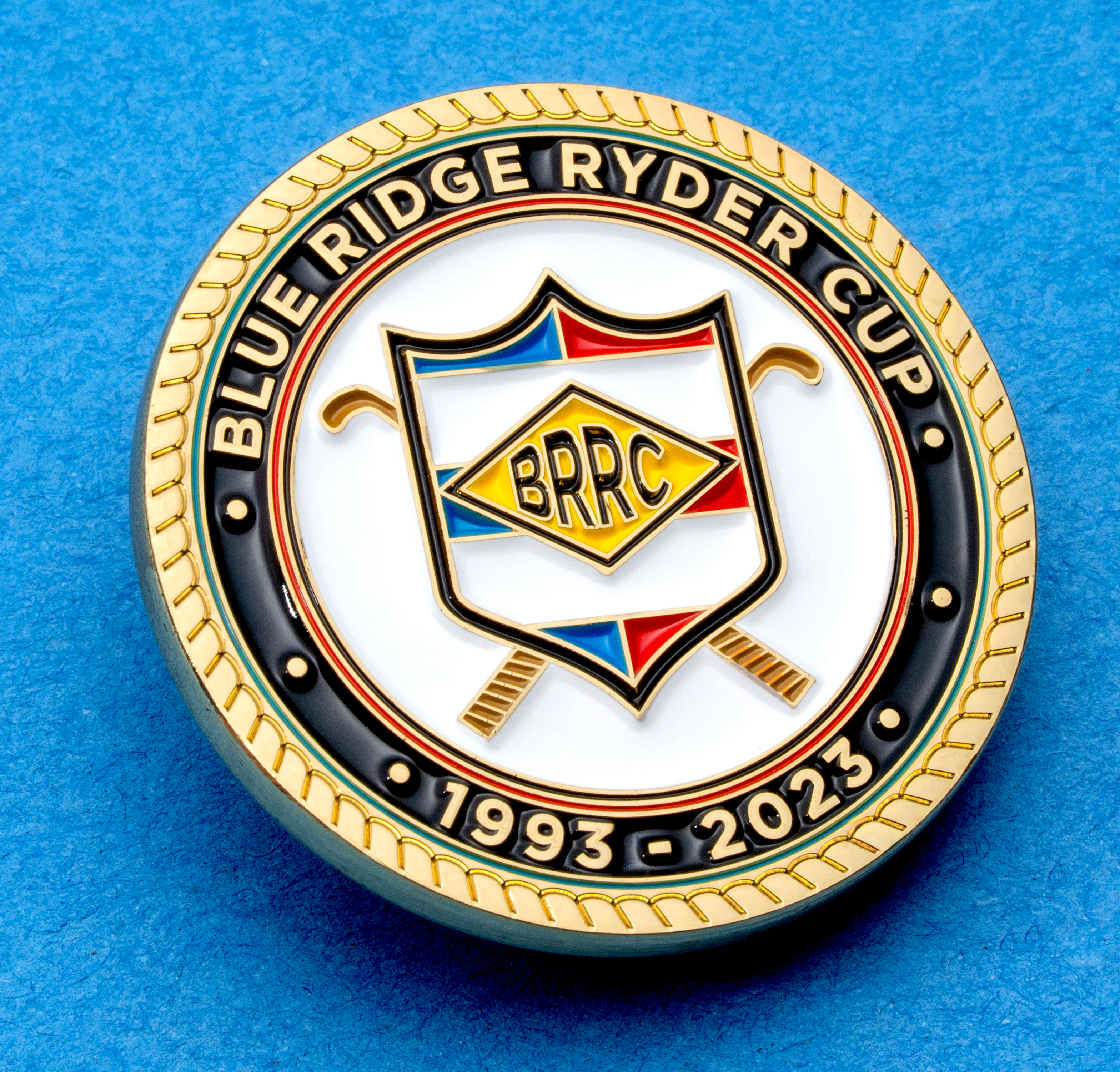 Circular Blue Ridge Ryder Cup challenge coin with a shield emblem, crossed golf clubs, and 1993-2023 dates on a blue background.