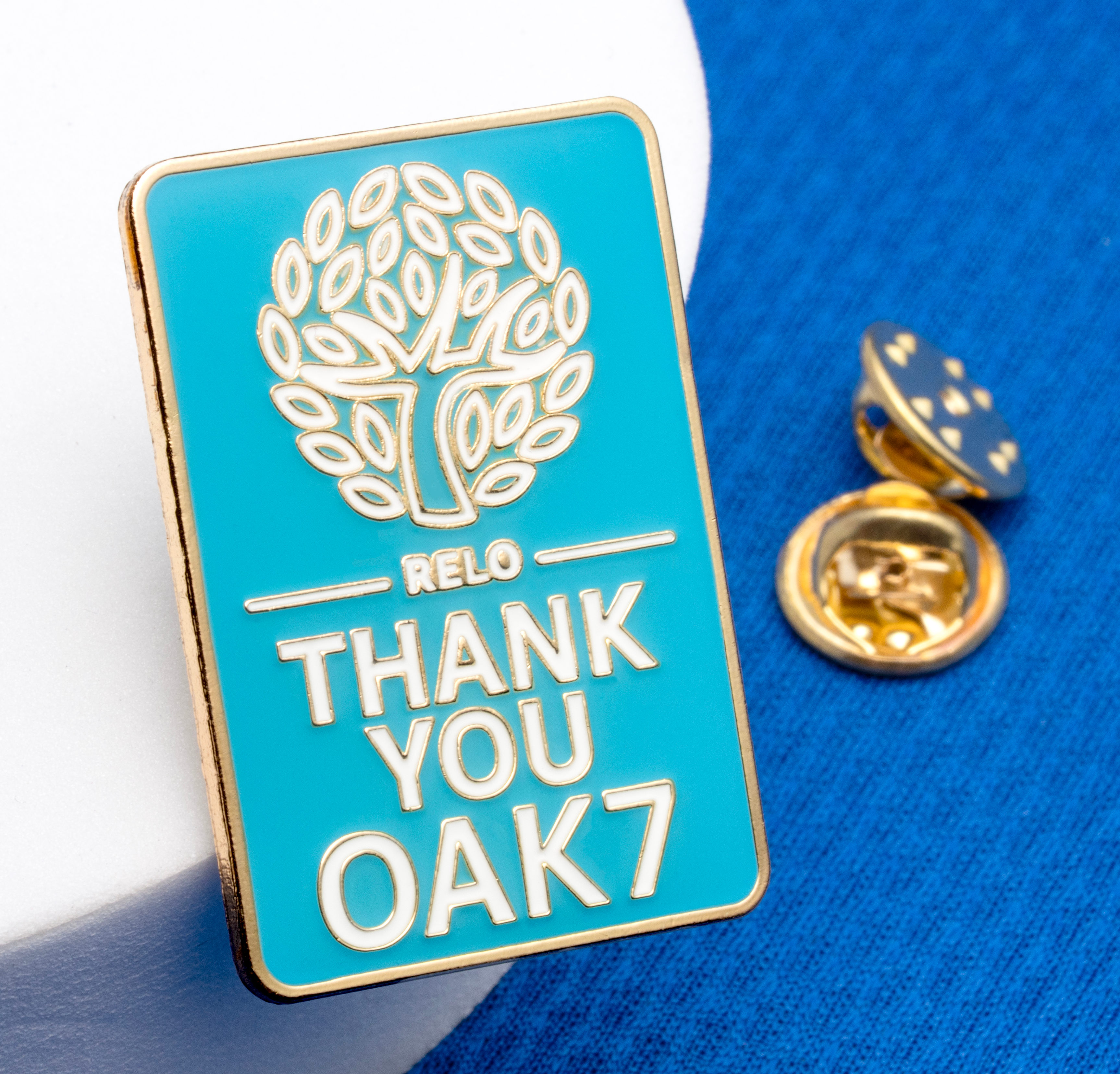 Rectangular teal lapel pin with a gold tree logo and the text RELO THANK YOU OAK7.