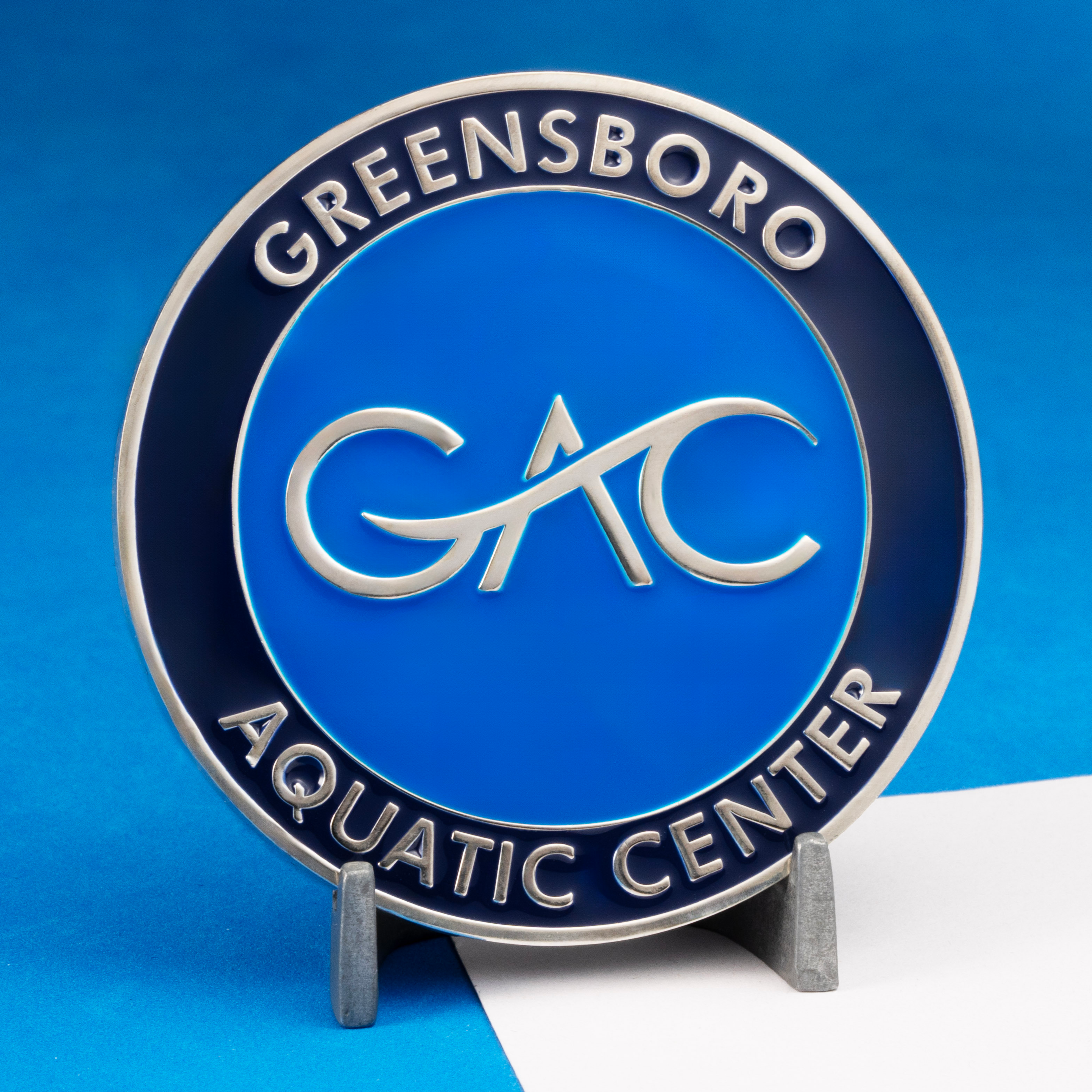 Circular challenge coin for the Greensboro Aquatic Center featuring a blue GAC logo in the center and a navy blue outer ring with silver text.