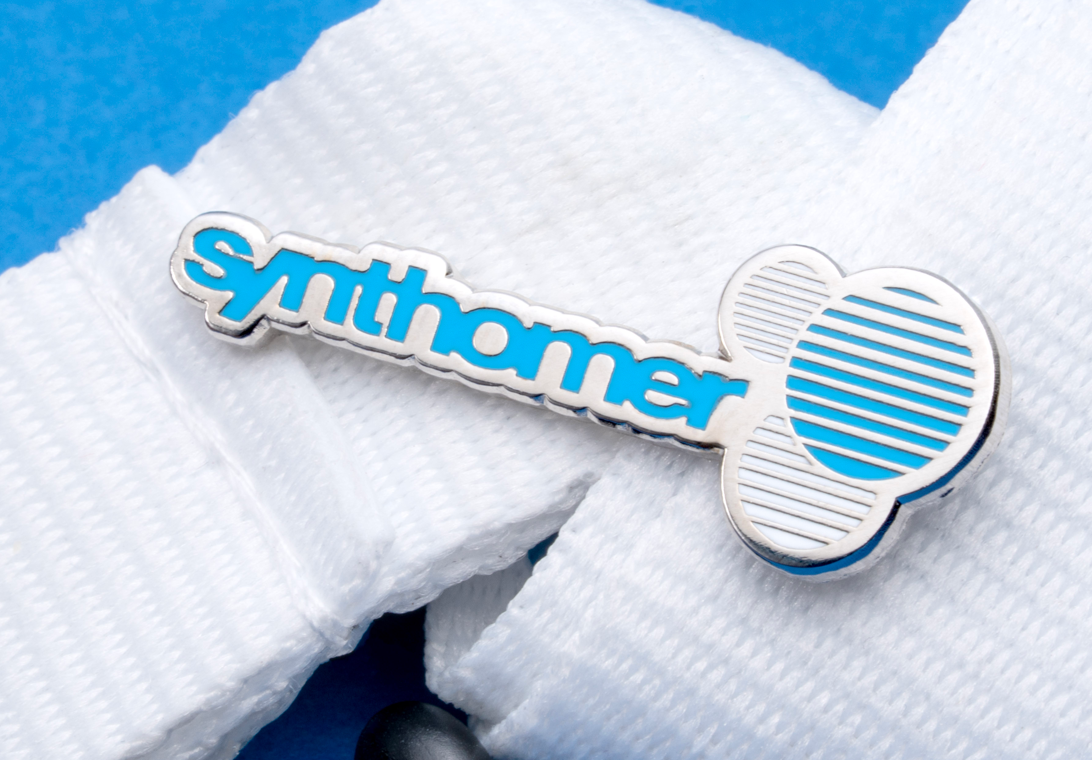 Synthomer brand lapel pin with blue enamel lettering and a silver circular logo on a white lanyard.