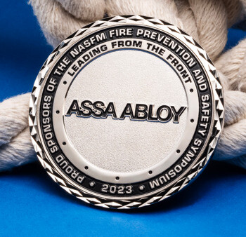 Silver ASSA ABLOY challenge coin featuring NASFM Fire Prevention and Safety Symposium text, displayed against a blue background with a white rope.