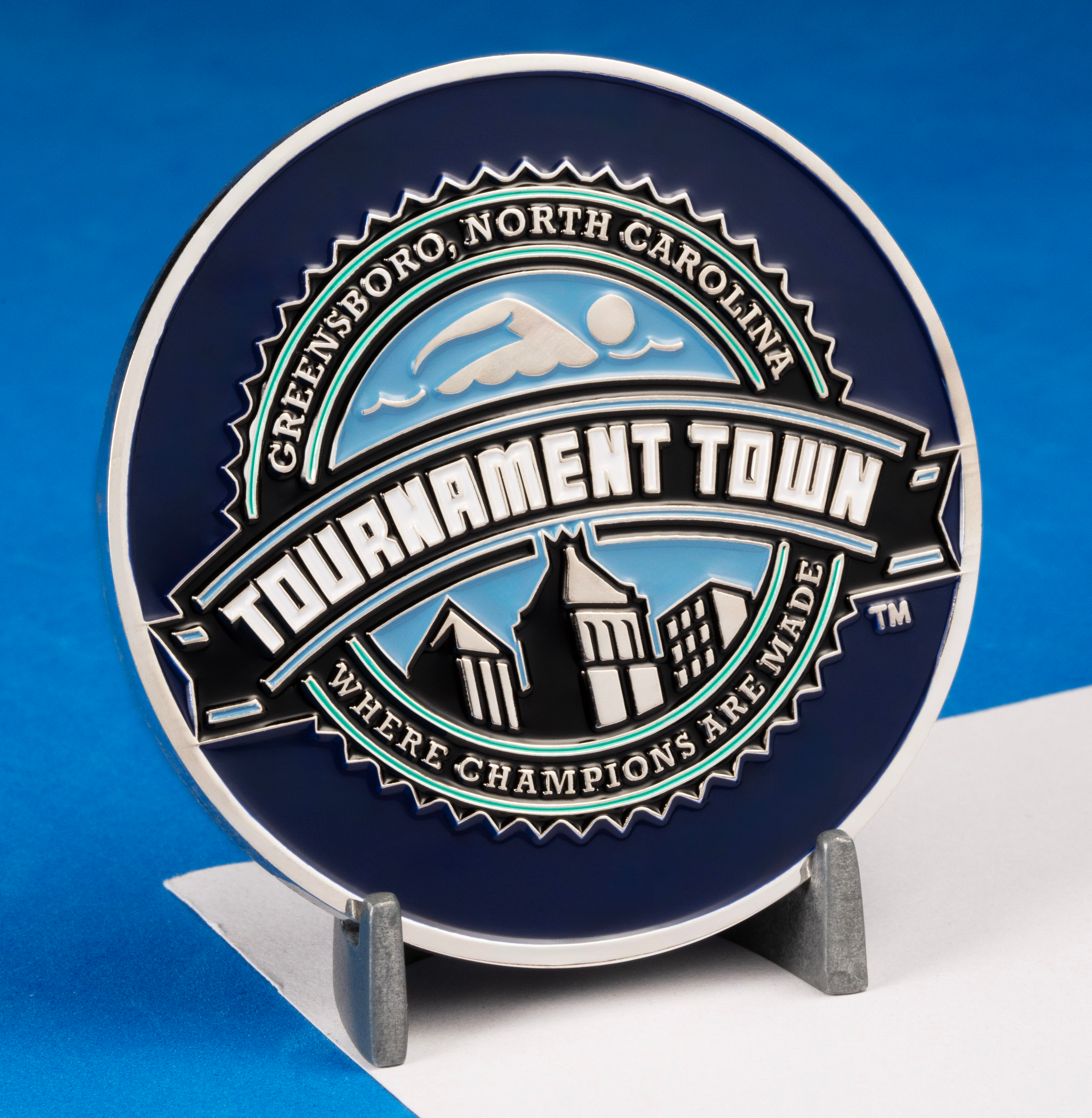 Circular blue and silver challenge coin featuring Tournament Town Greensboro North Carolina text with a swimmer icon and city skyline illustration.