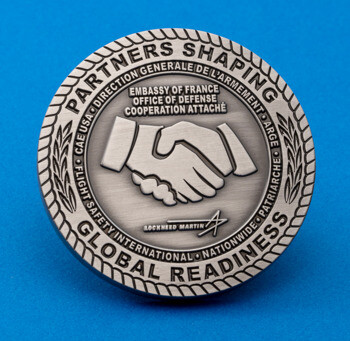 Antiqued silver challenge coin featuring a handshake, Embassy of France Office of Defense insignia, and Lockheed Martin logo on a blue background.