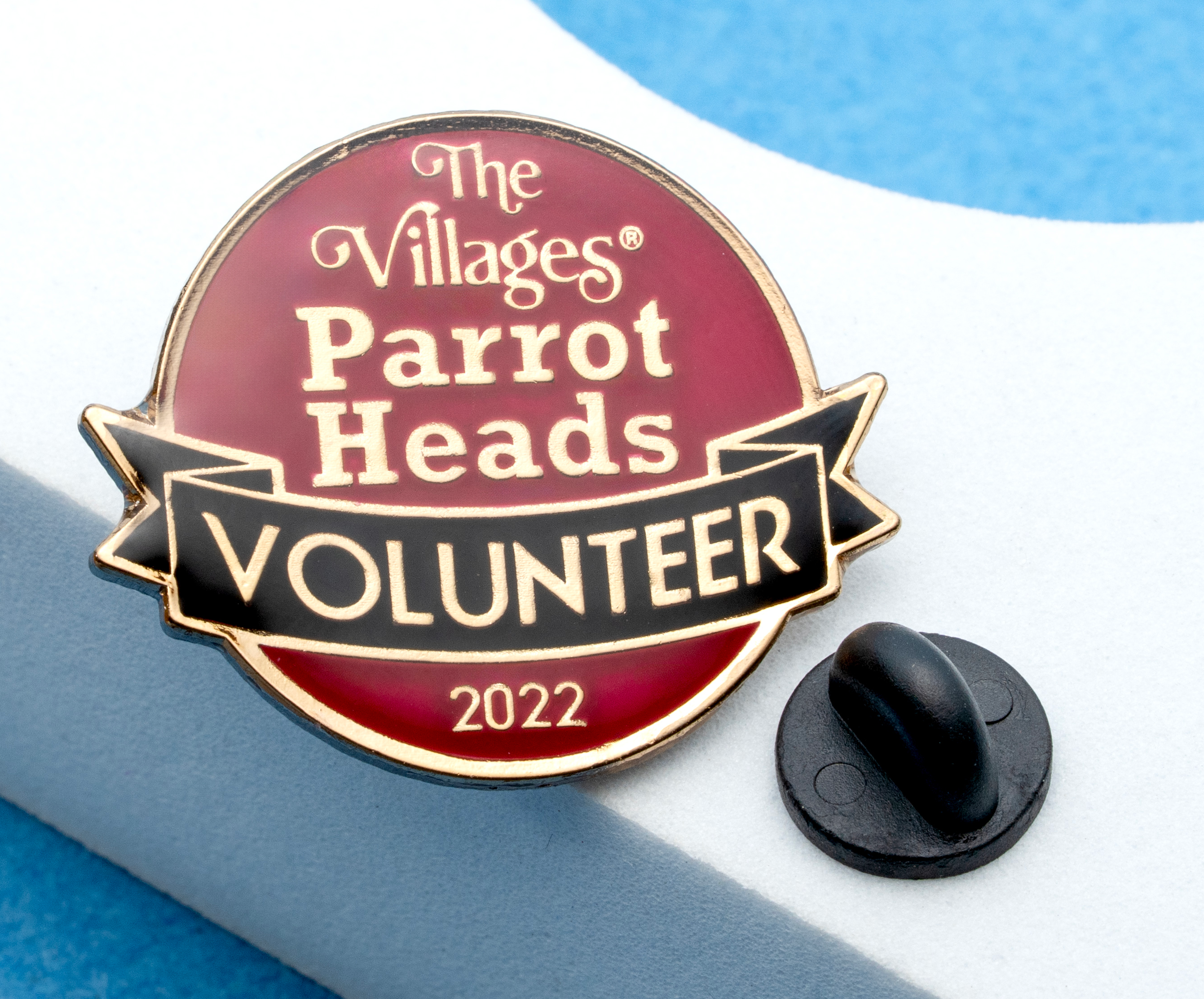 Round enamel lapel pin with maroon and black sections for The Villages Parrot Heads Volunteer 2022 with a black rubber clutch.