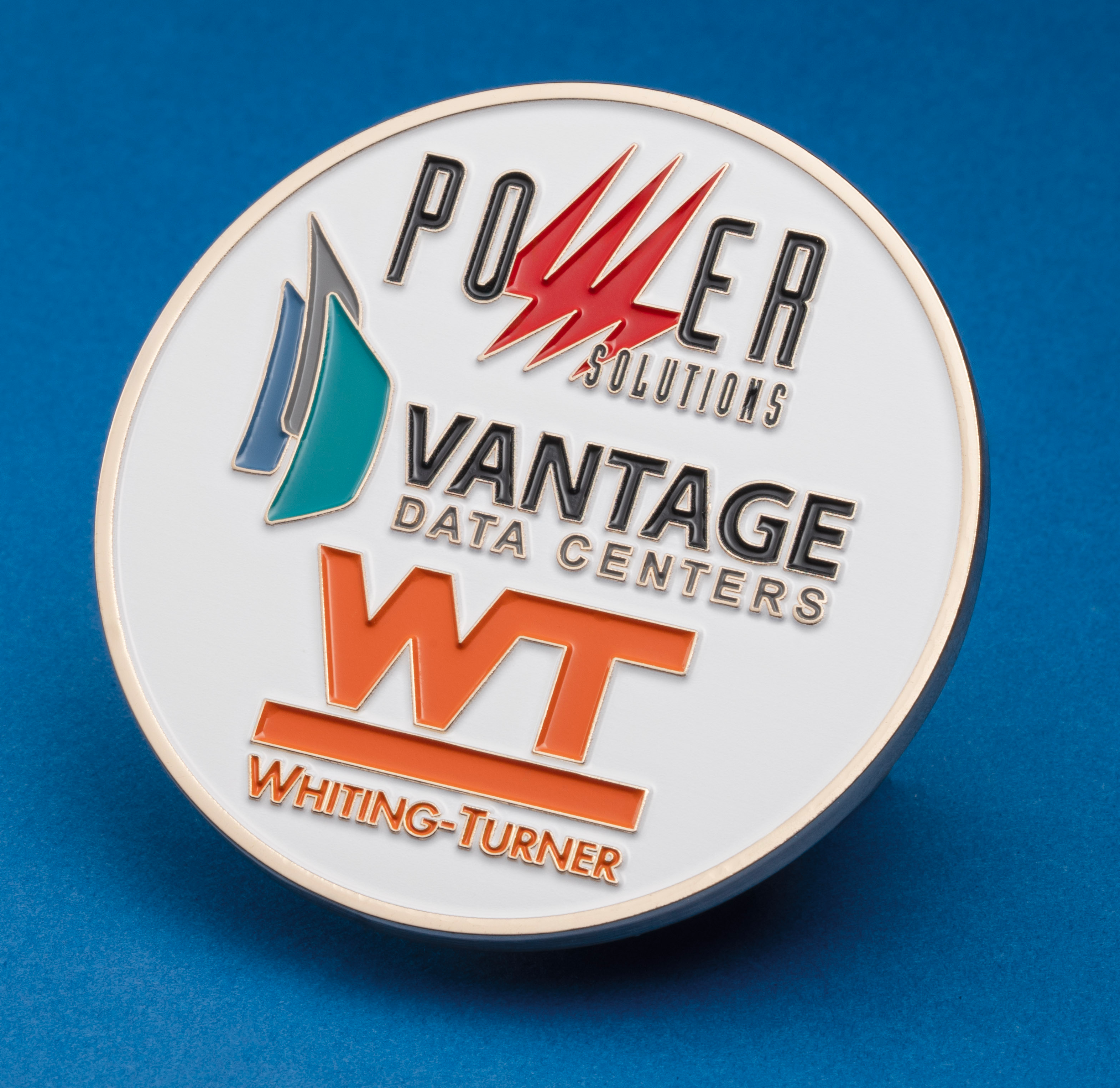 Round white enamel challenge coin featuring logos for Power Solutions, Vantage Data Centers, and Whiting-Turner on a blue background.