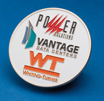 Round white enamel challenge coin featuring logos for Power Solutions, Vantage Data Centers, and Whiting-Turner on a blue background.