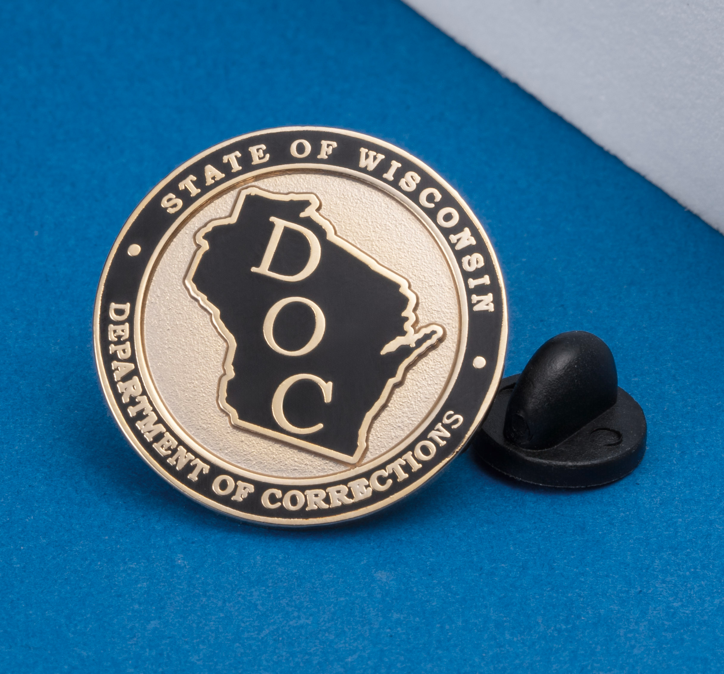 Circular lapel pin for the State of Wisconsin Department of Corrections featuring a black outline of the state with DOC lettering on a gold background.