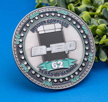 Antiqued silver challenge coin with 'M-130' and '62 Years of Service' text against a blue background and green foliage.