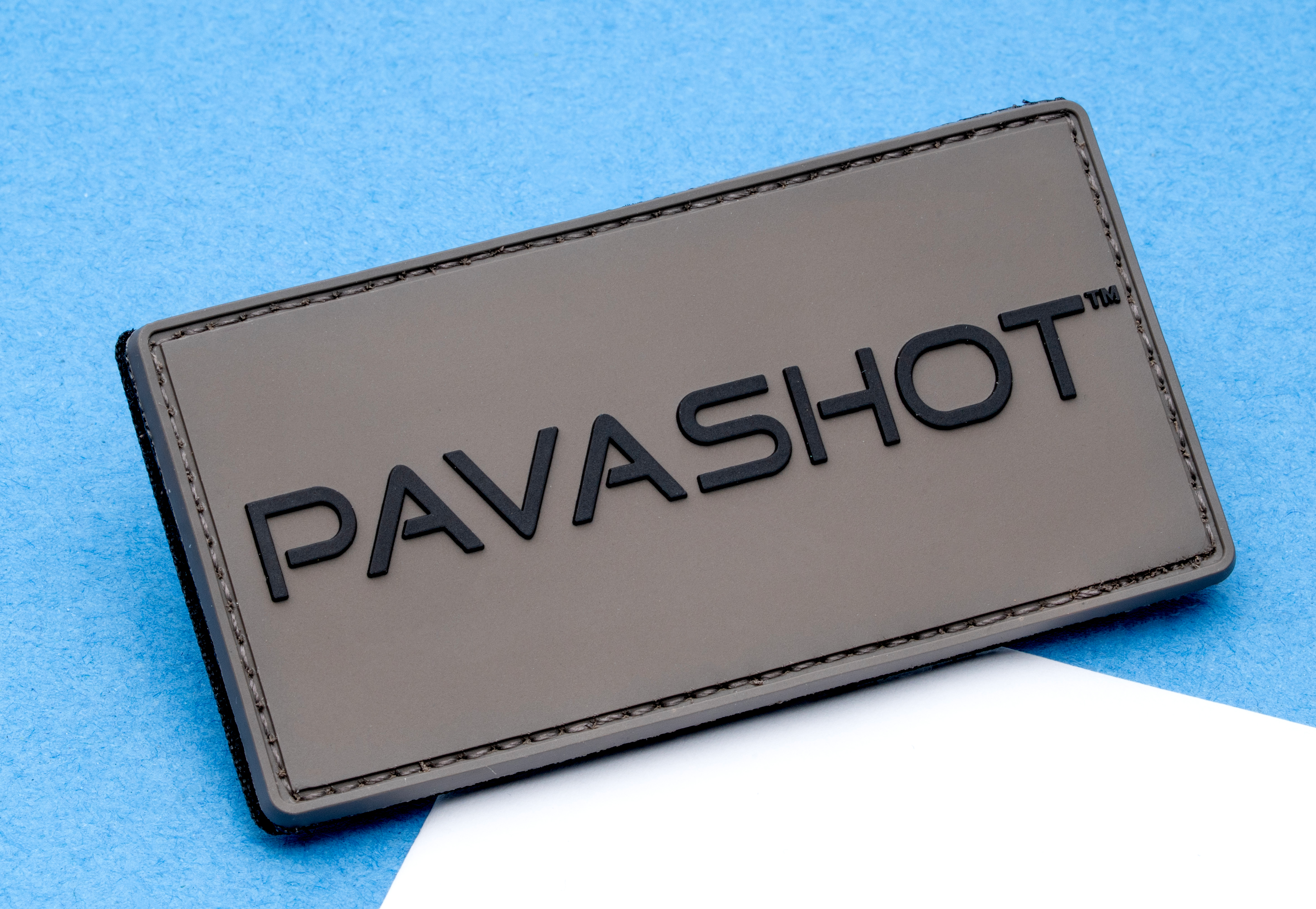 Rectangular grey PVC patch with black PAVASHOT text in raised lettering and stitched border on a blue background.