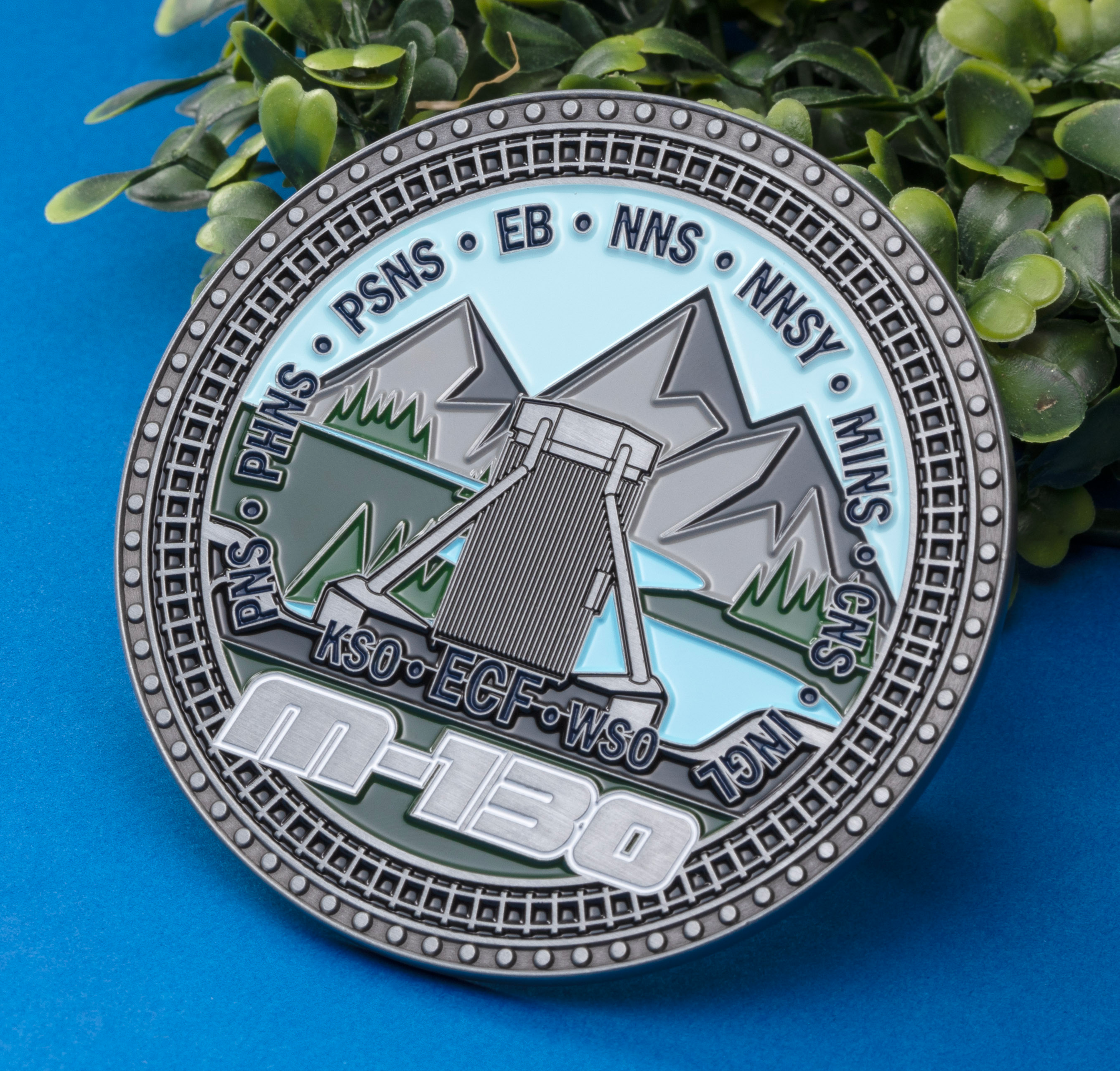 Circular challenge coin featuring an M-130 design with mountains, water, and industrial acronyms including PHNS, PSNS, and EB against a blue background.
