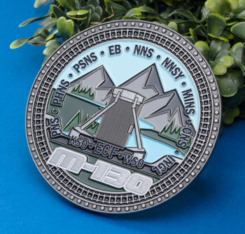 Circular challenge coin featuring an M-130 design with mountains, water, and industrial acronyms including PHNS, PSNS, and EB against a blue background.