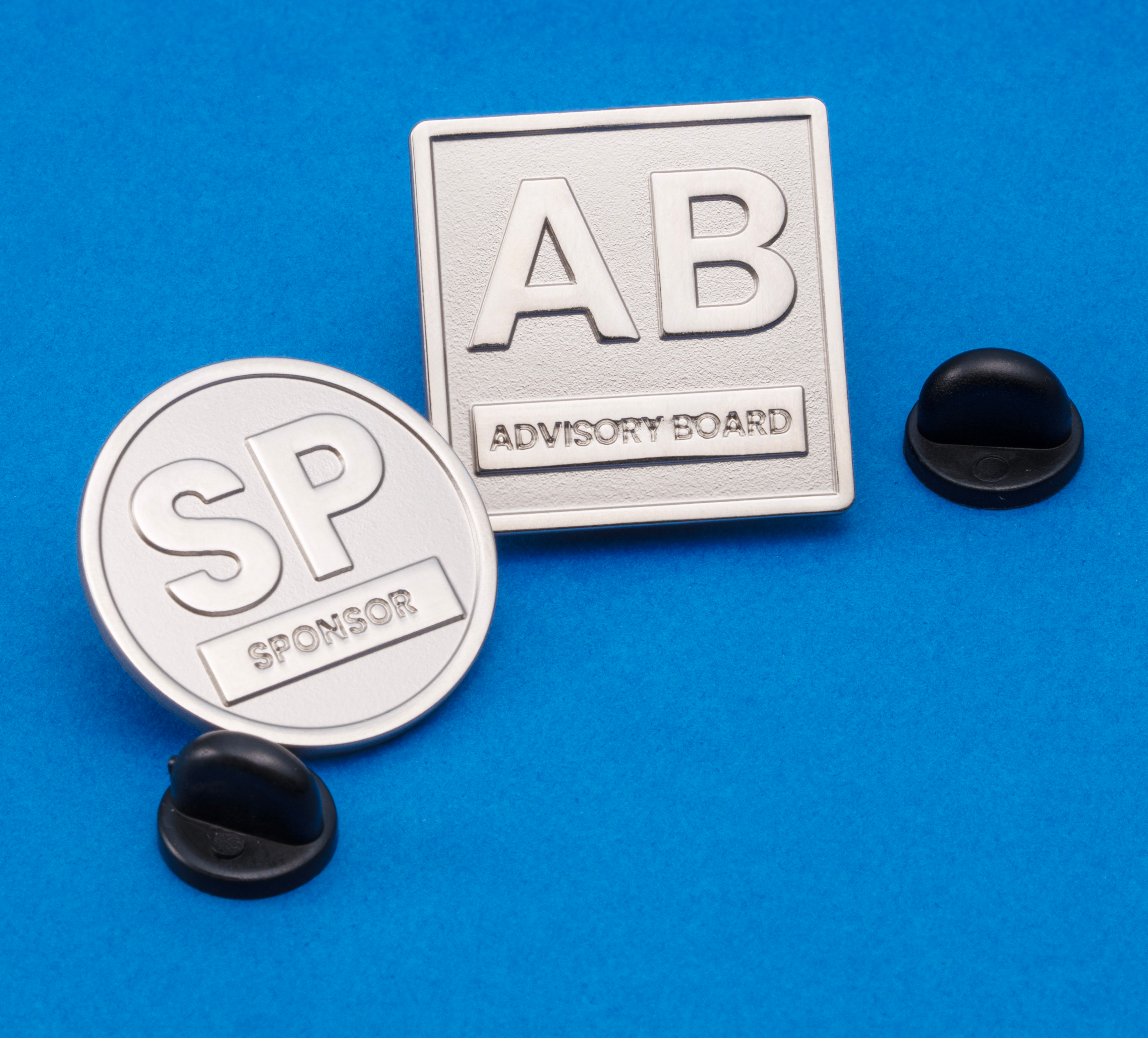 A round silver pin with SP Sponsor text and a square silver pin with AB Advisory Board text, both with black rubber clutches on a blue background.