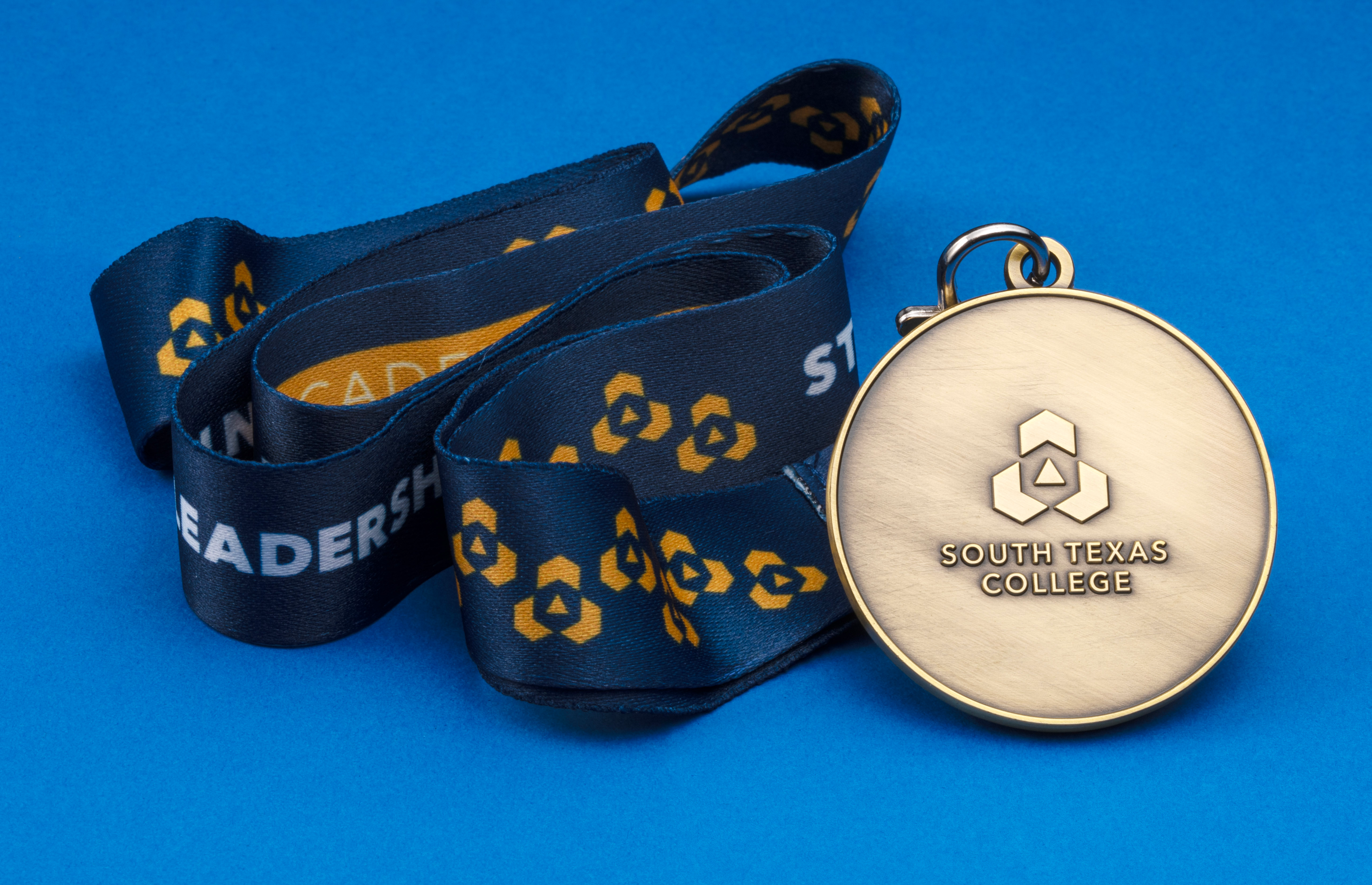 Antique brass medallion featuring the South Texas College logo and text, accompanied by a navy blue and gold printed ribbon on a blue background.