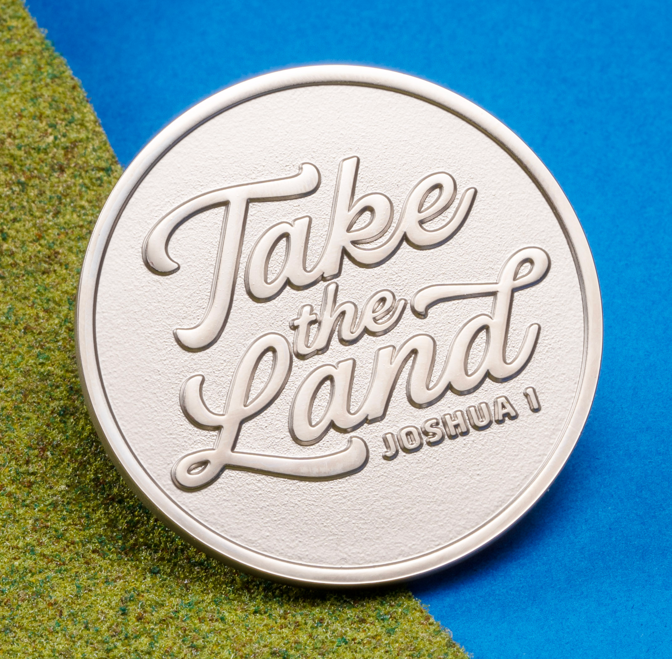 A circular silver coin featuring the embossed text 'Take the Land' and 'Joshua 1' resting on a green and blue background.