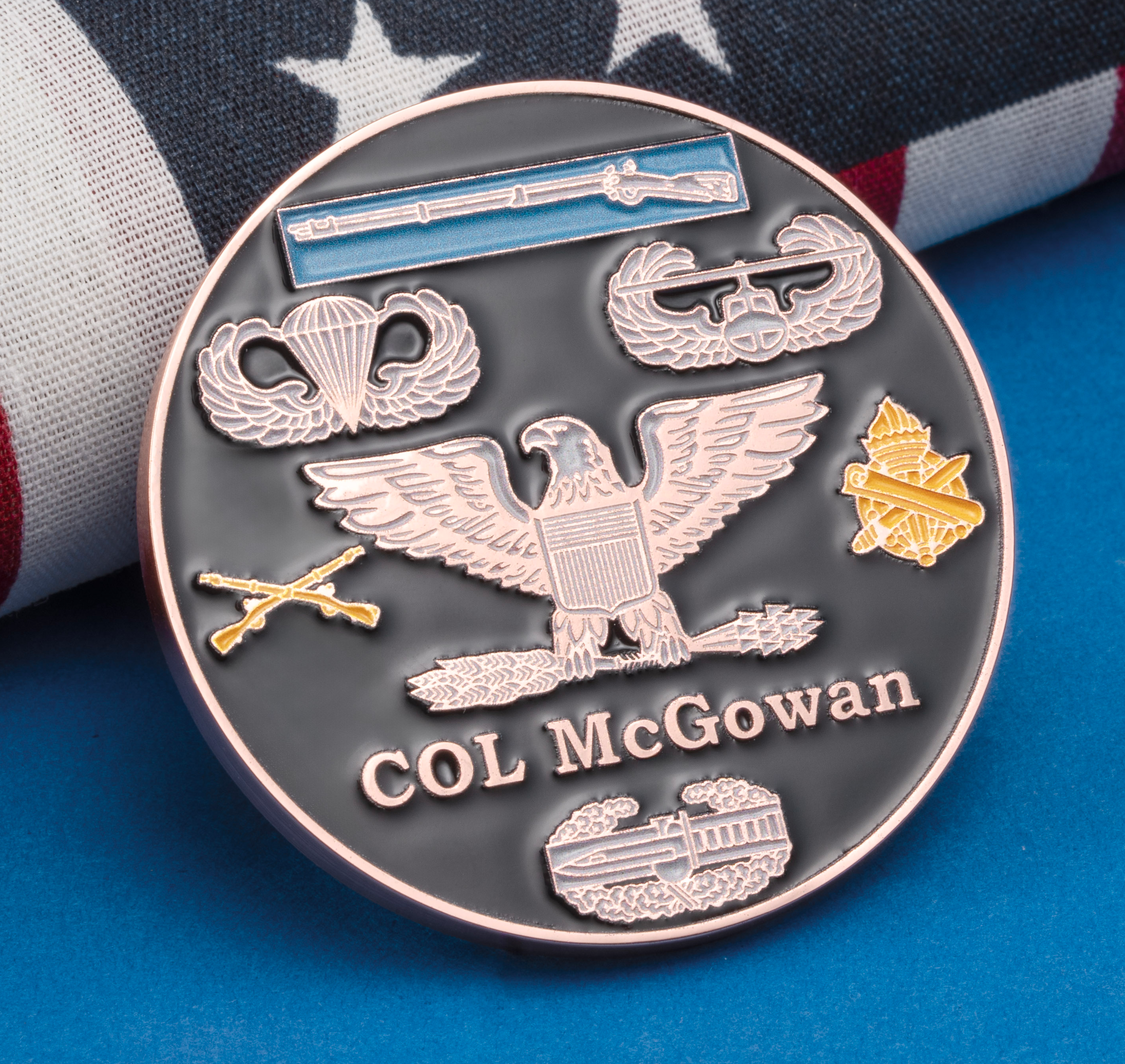 A black and rose gold challenge coin featuring military badges, an eagle, and the name COL McGowan, resting in front of an American flag.