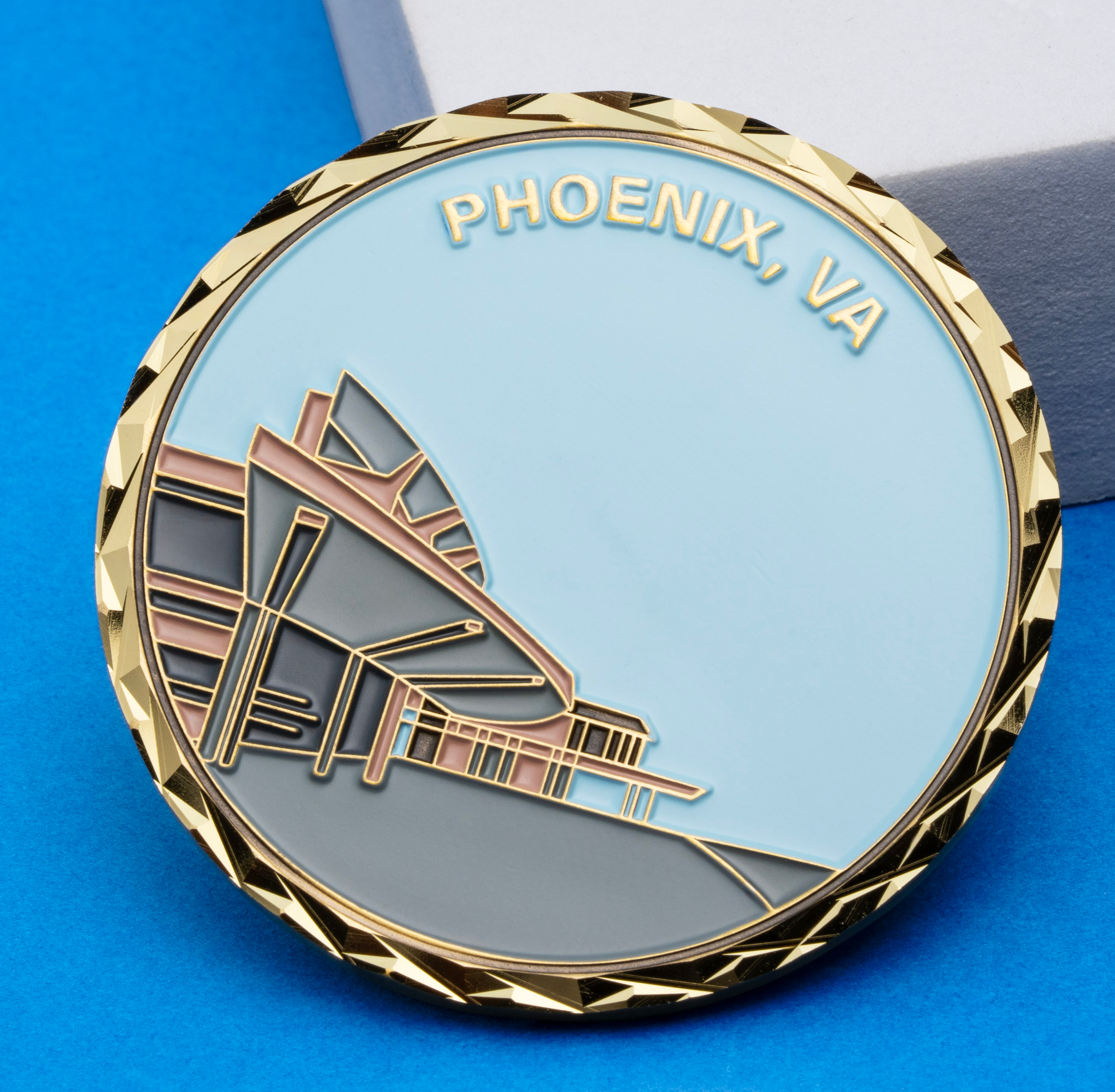 Circular gold challenge coin with a diamond-cut edge featuring a modern building illustration and the text Phoenix, VA.