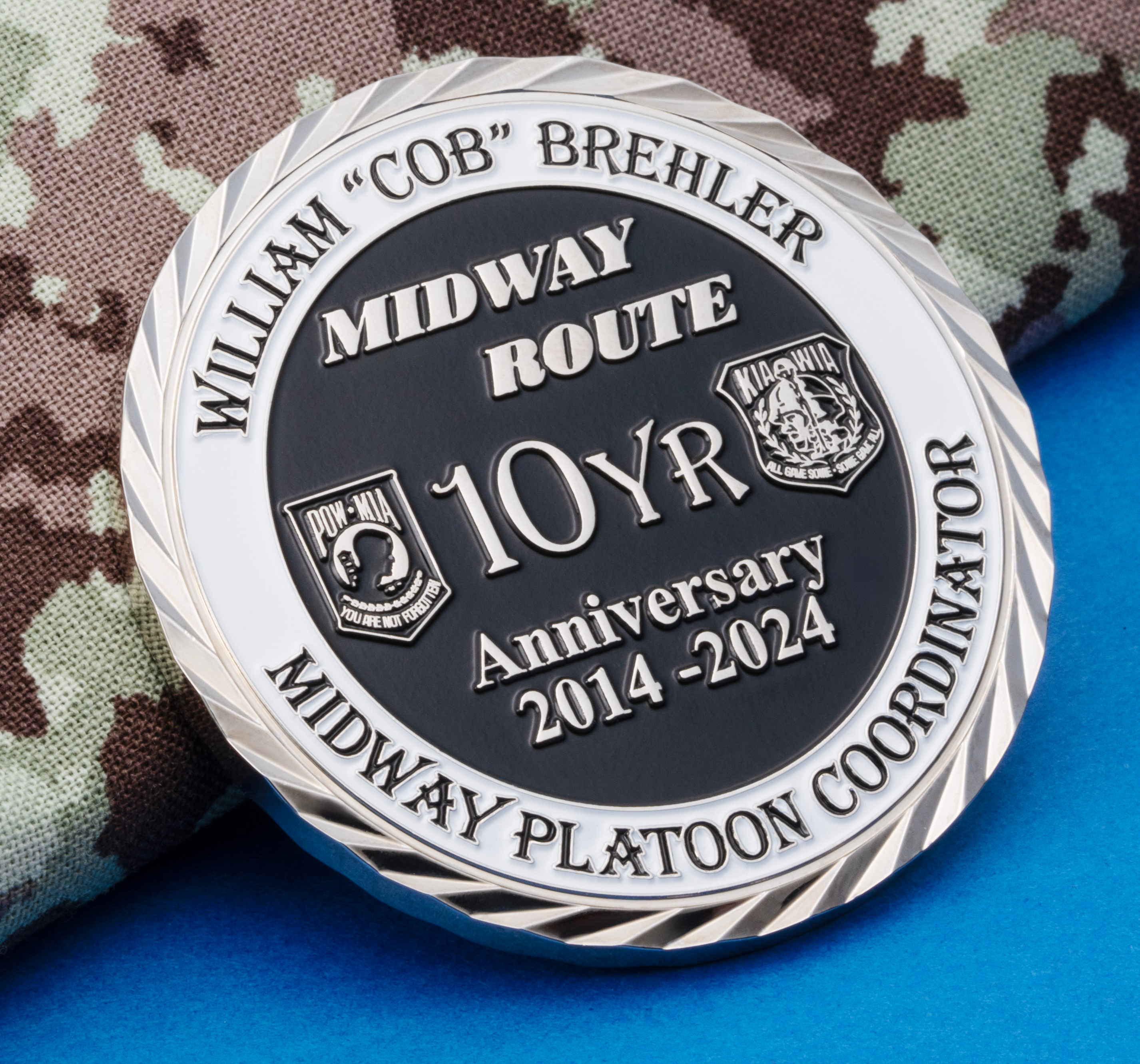 Silver challenge coin with black recessed background commemorating the 10 Year Anniversary of the Midway Route Midway Platoon Coordinator on camouflage fabric.