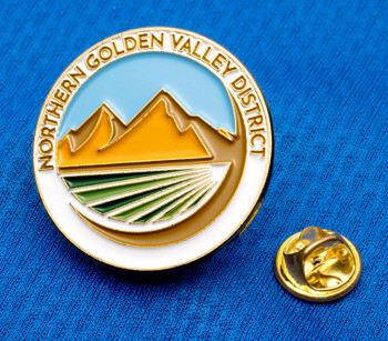 A circular enamel lapel pin for Northern Golden Valley District featuring gold mountains, green fields, and a light blue sky on a blue fabric background.