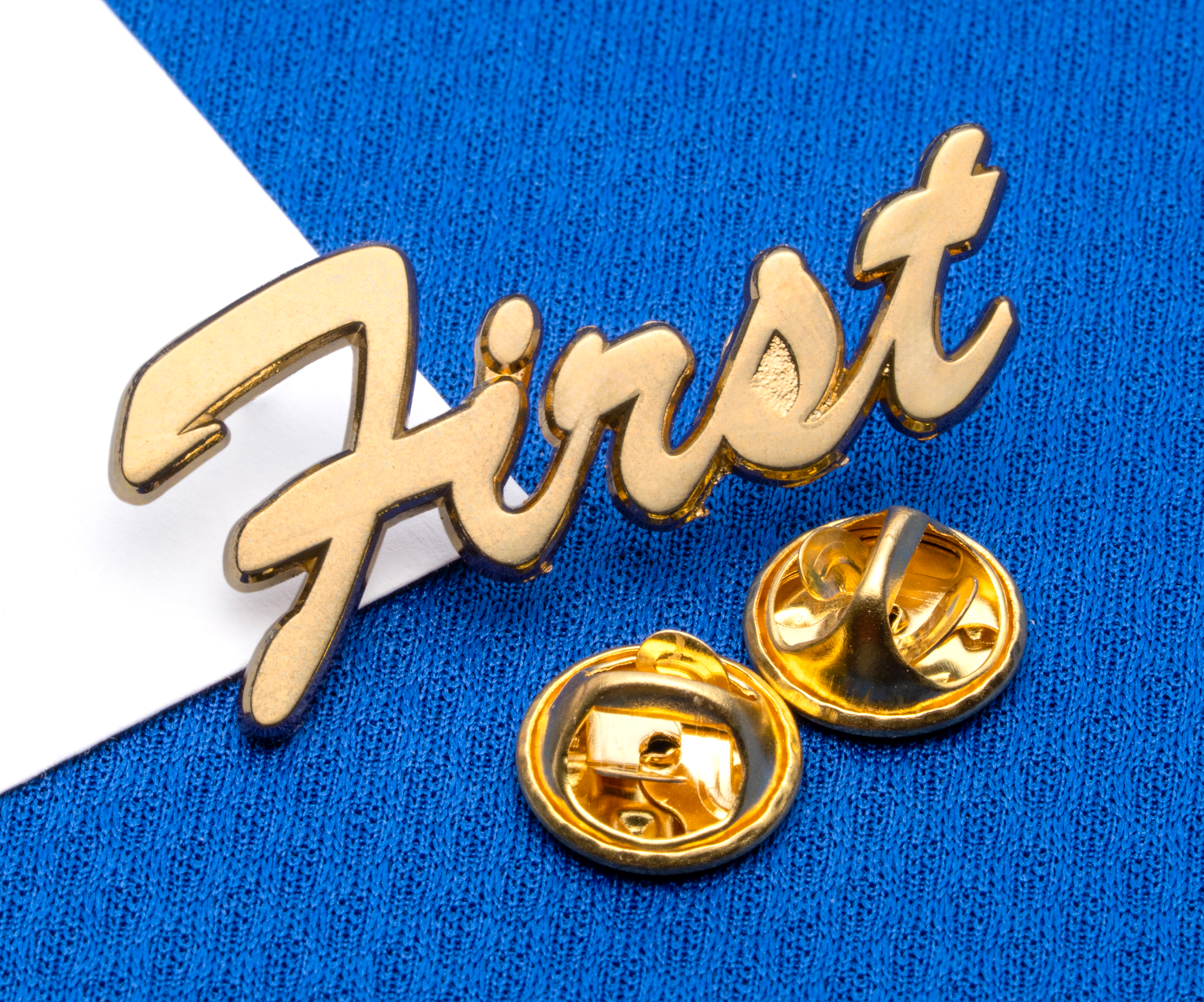 Gold script lapel pin reading First with two butterfly clutch backings on a blue textured background.
